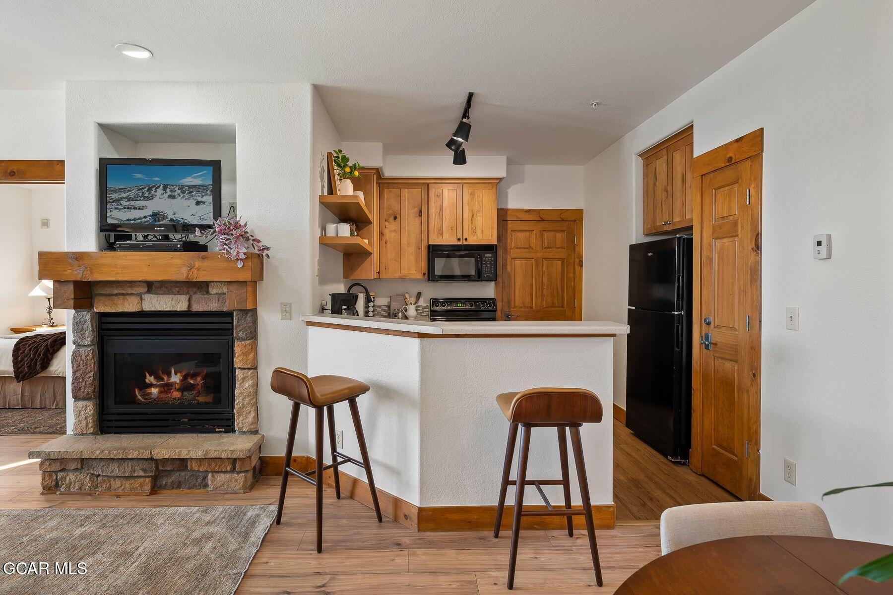 Open-plan condo kitchen and living area with wood cabinetry, breakfast bar, and stone fireplace.