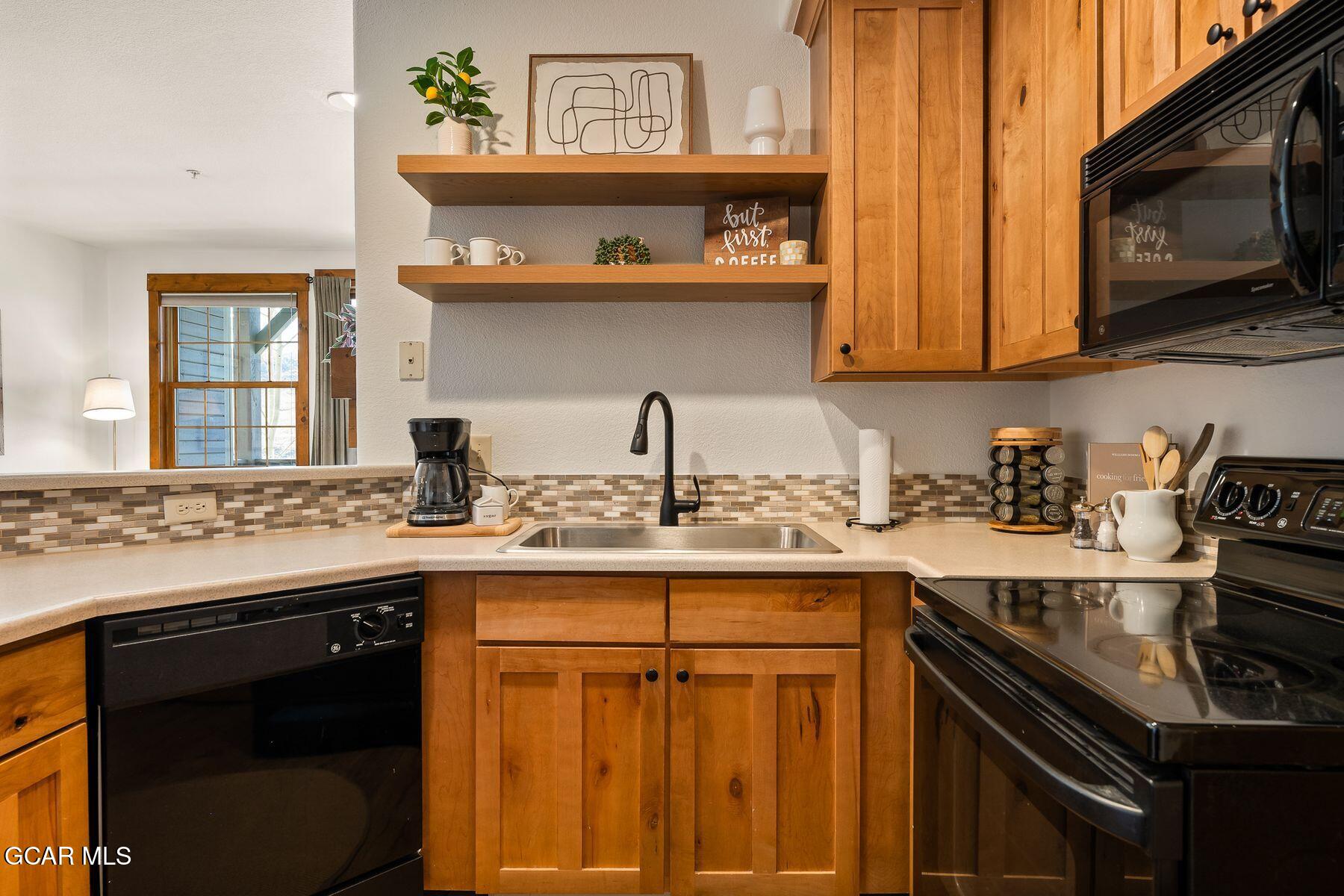Kitchen with warm wood cabinets, mosaic backsplash, open shelves, and black appliances.