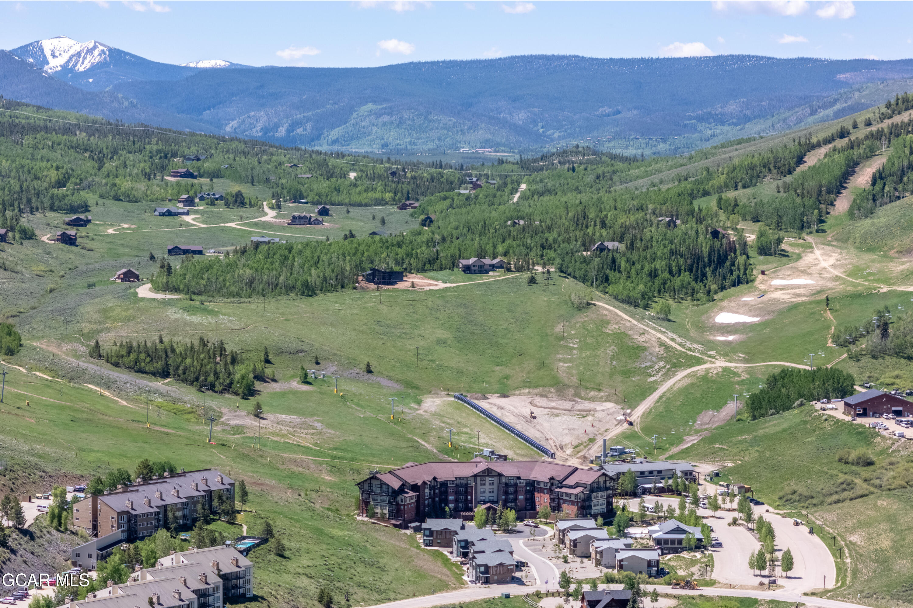 Aerial view of resort condos below a green hillside and distant snow-capped mountains.