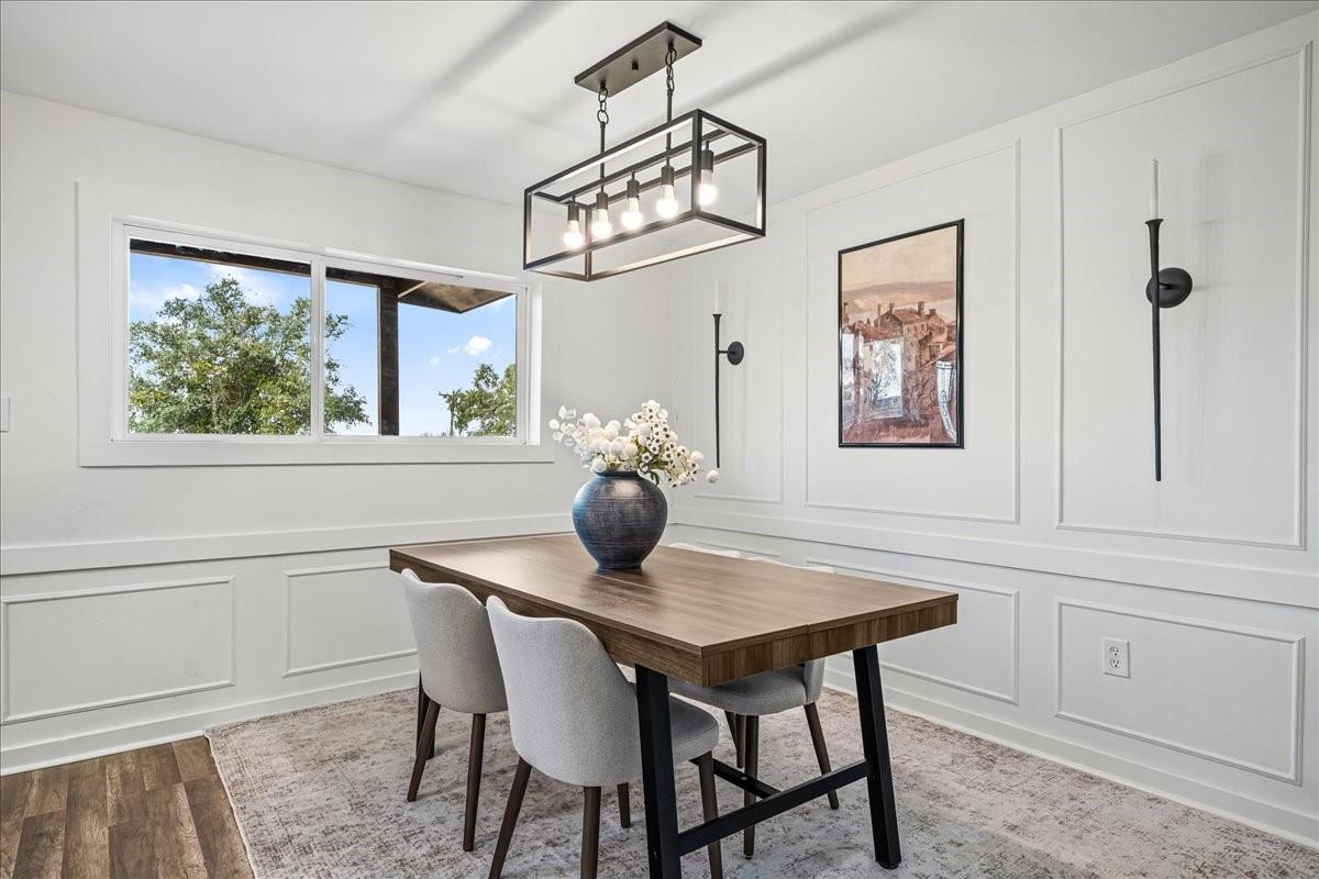 Dining room with a wood table, upholstered chairs, white paneled walls, and a wide window.