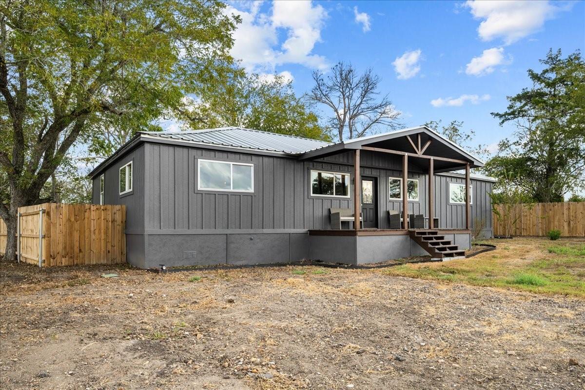 Single-story gray house with a metal roof, covered porch, and fenced gravel yard.
