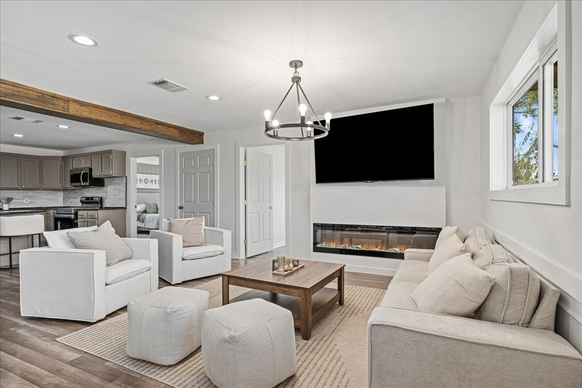Staged living room with a sectional, linear fireplace, wall-mounted TV, and exposed beam overlooking the kitchen.