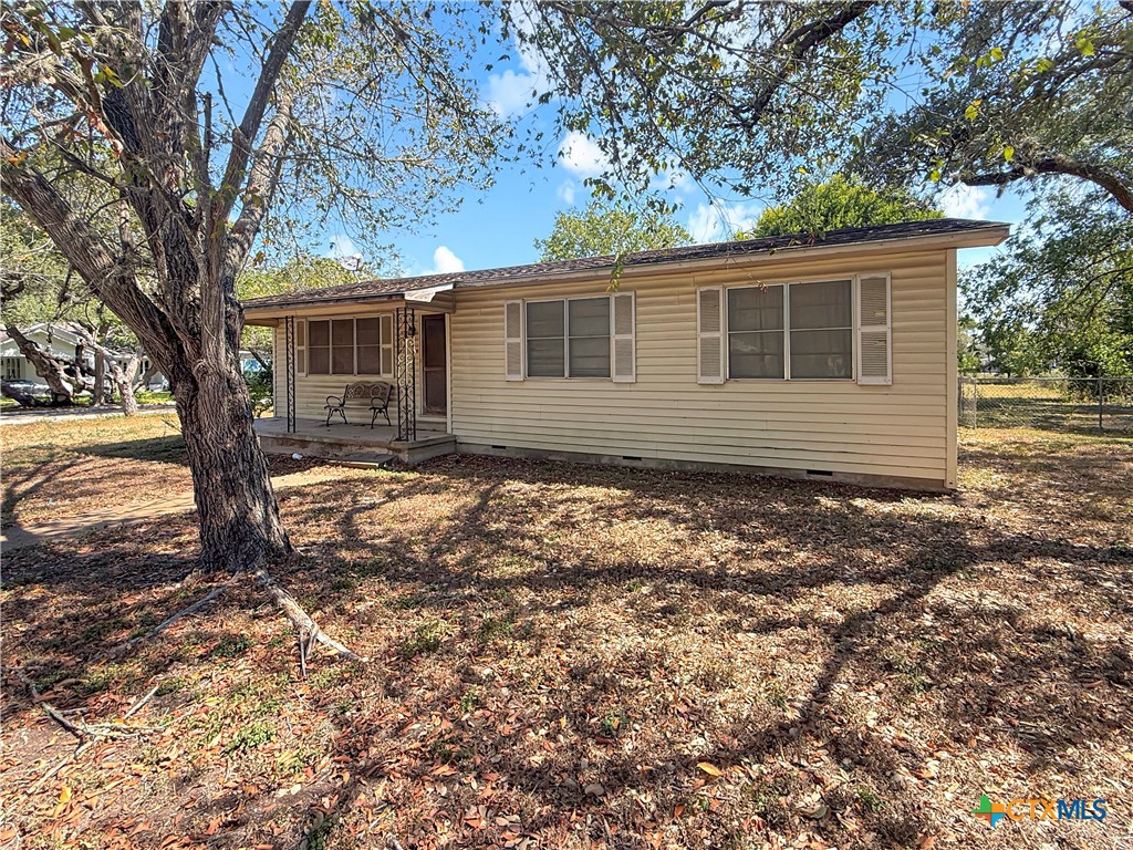Single-story beige house with a covered porch beneath large shade trees.