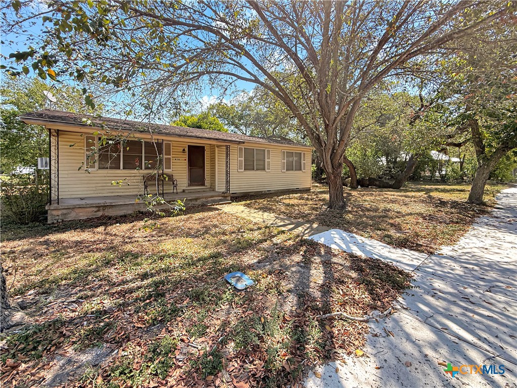Single-story beige house beneath large shade trees, with a small covered front porch.