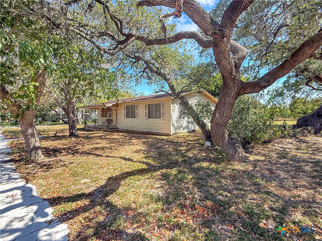 Single-story light-colored house with a covered porch beneath mature oak trees.