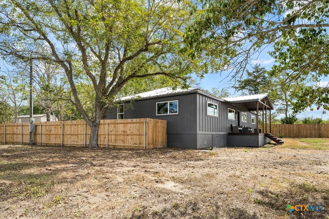 Dark gray single-story home with a covered porch, wooden privacy fence, and large shade tree.
