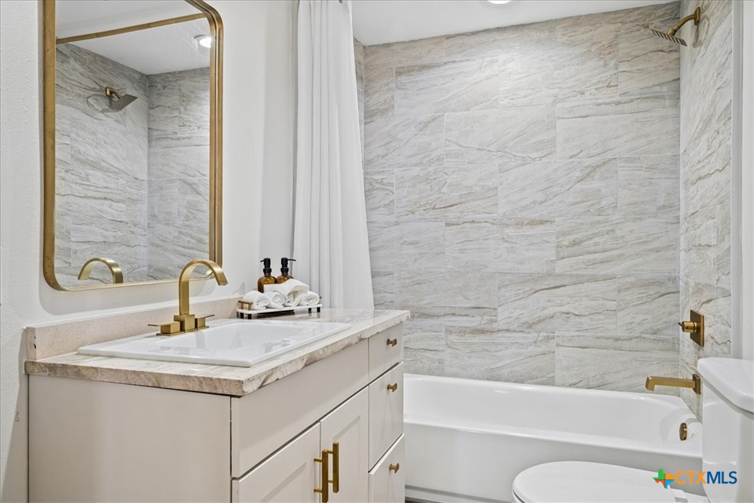Bathroom with a white vanity, gold-framed mirror, and tub-shower lined with light veined tile.