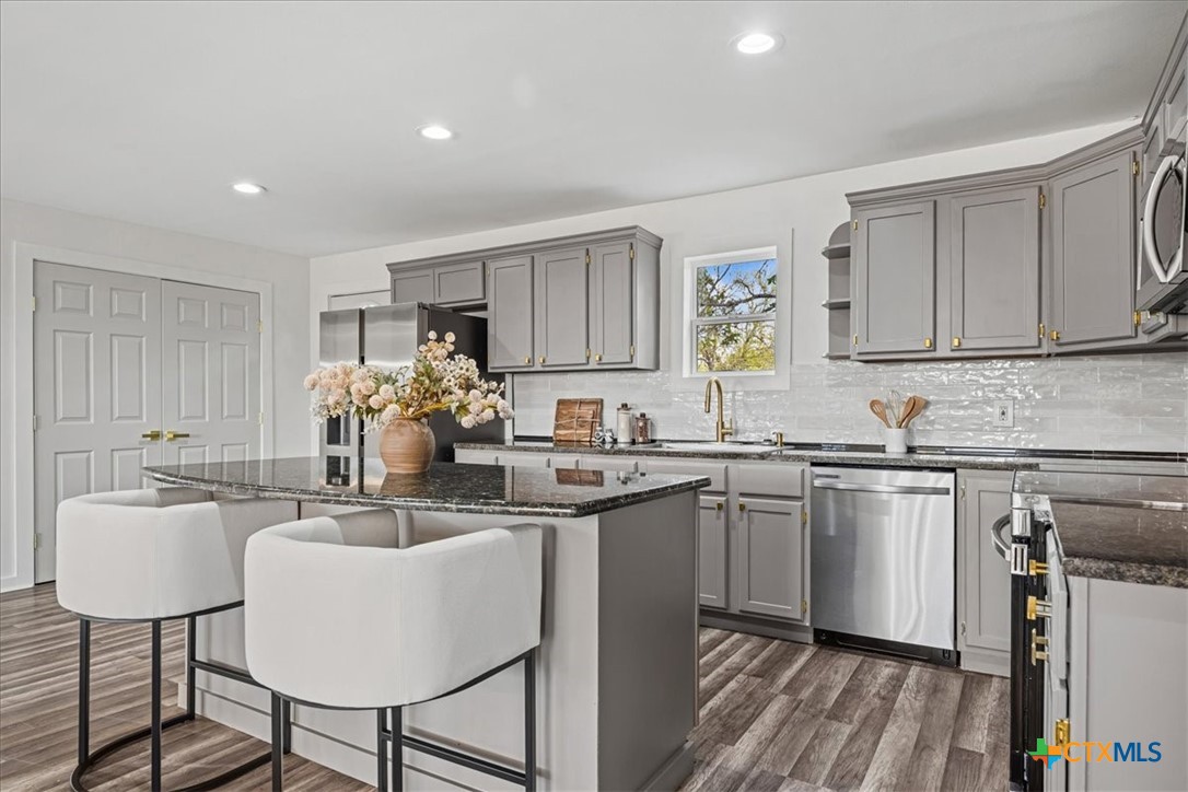 Staged kitchen with gray cabinets, a dark stone island, two white stools, and stainless steel appliances.