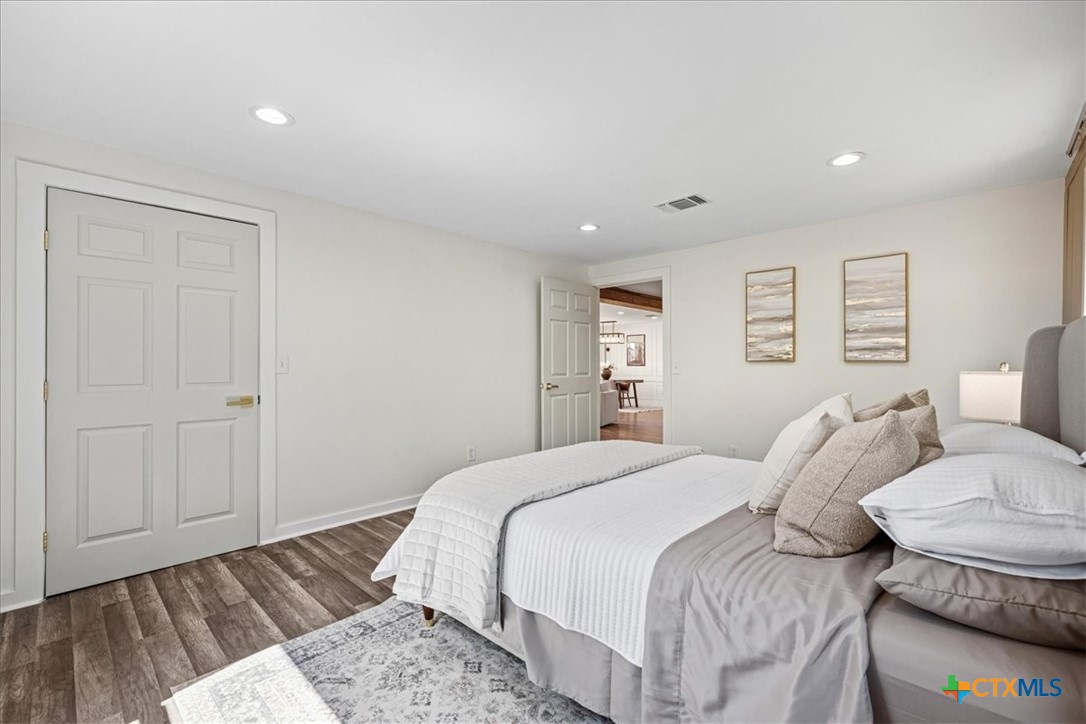 Staged bedroom with a large white-and-gray bed, patterned rug, and dark wood-look flooring.
