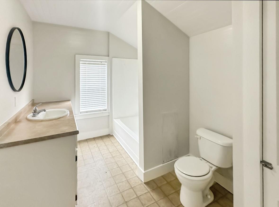 Bathroom with a bathtub behind a partition, toilet, and tan-countertop vanity beside a window.