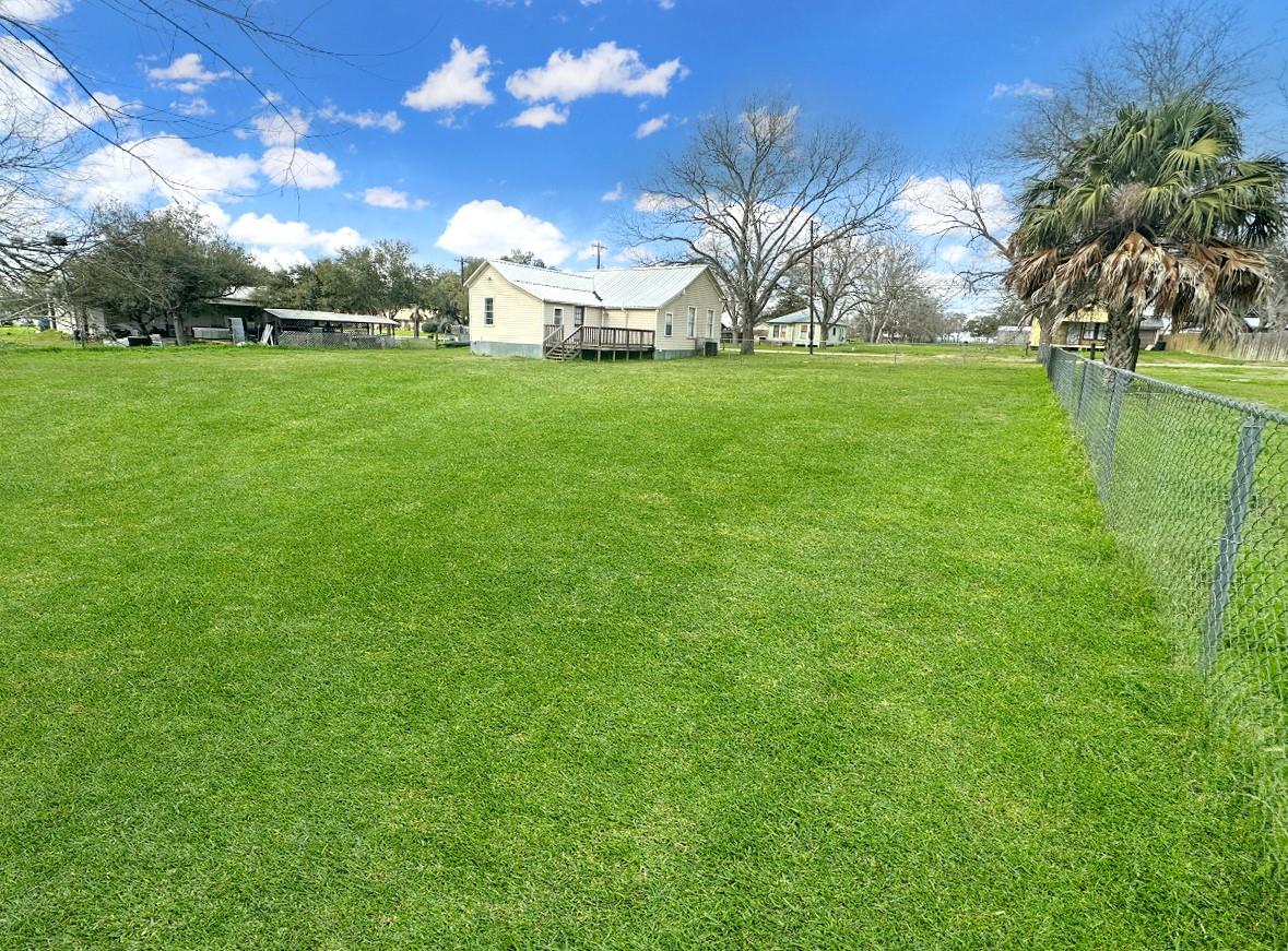 Small light-colored house beyond a broad lawn, with a chain-link fence and palm tree.