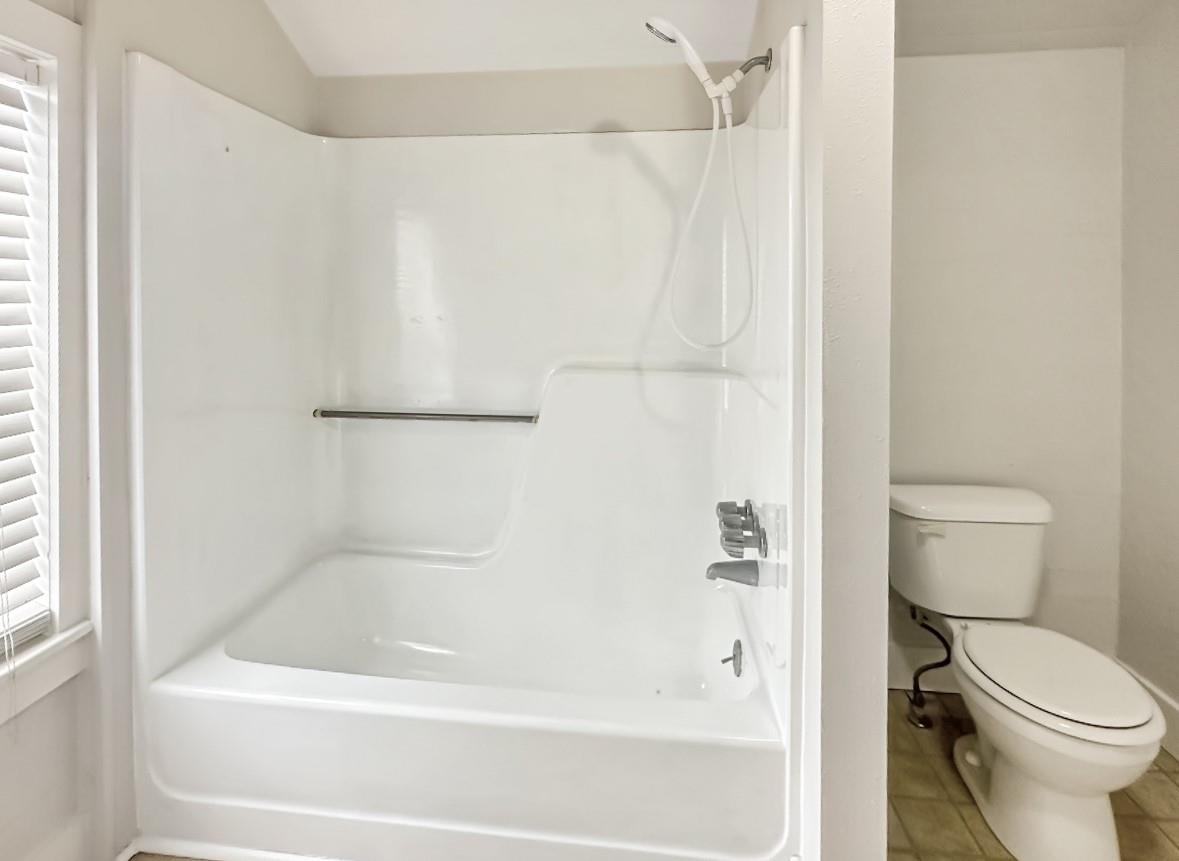 Bathroom with a white tub-shower and grab bar beside a toilet and window.