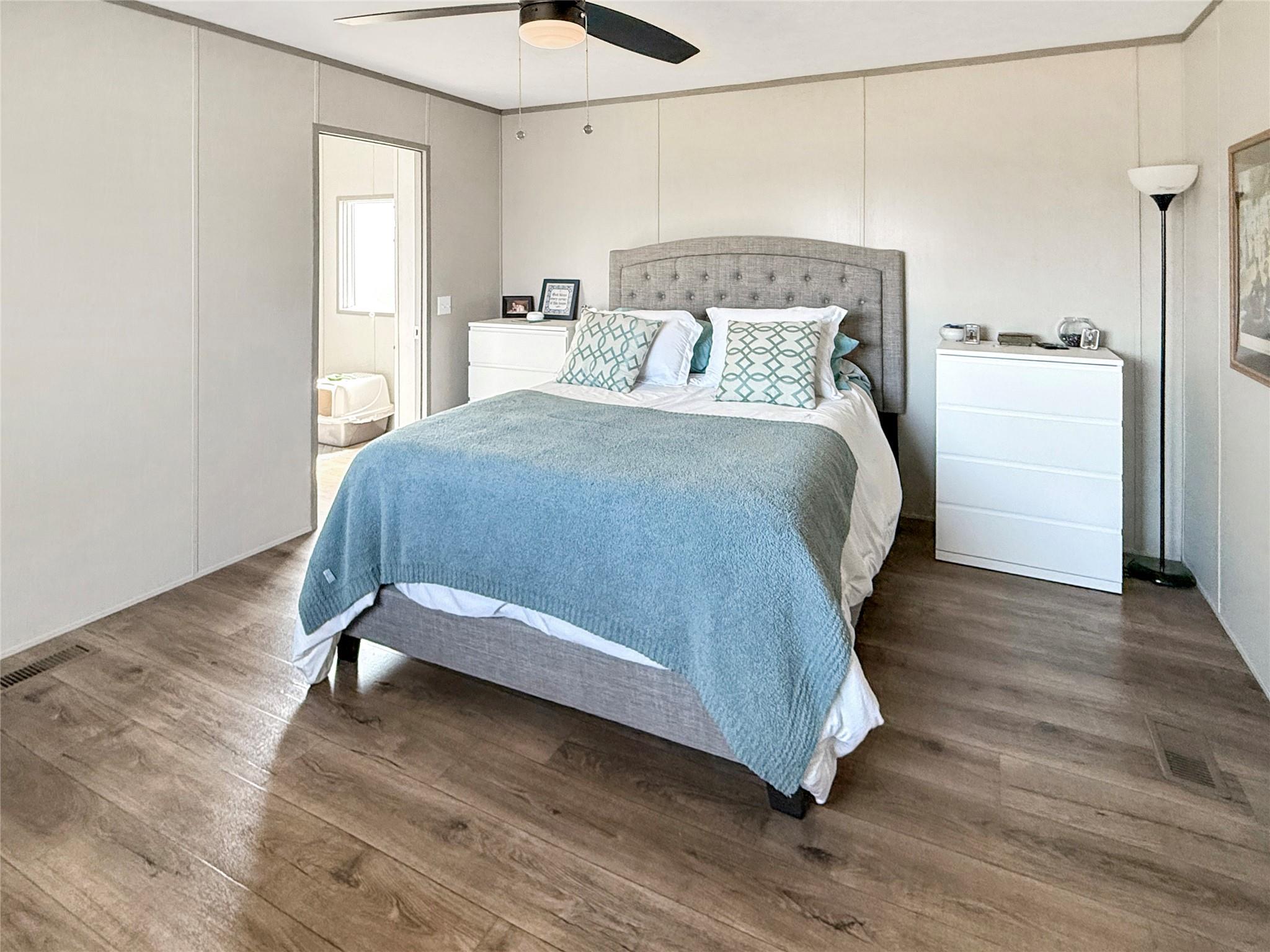 Staged bedroom with a blue-covered bed, gray tufted headboard, white dressers, and wood-look flooring.
