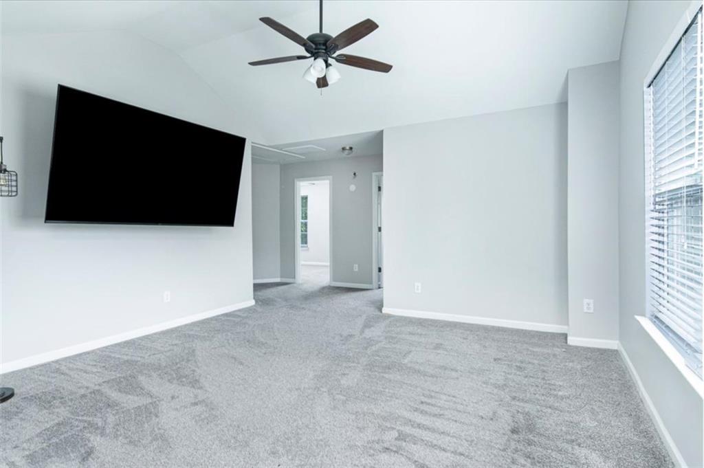 Empty carpeted bedroom with a ceiling fan, wall-mounted TV, and large window with blinds.
