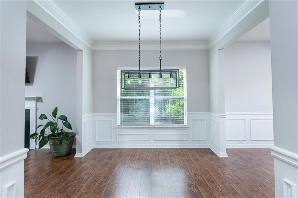 Empty dining room with dark hardwood floors, white wainscoting, and a large window beneath a hanging light.