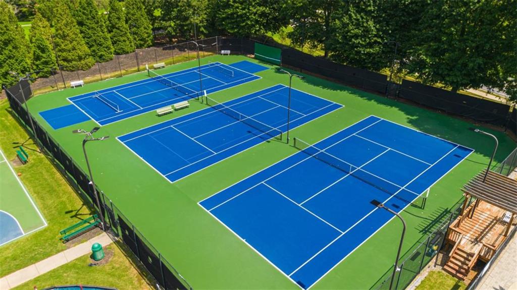 Several blue tennis courts with green surrounds, enclosed by fencing and trees.