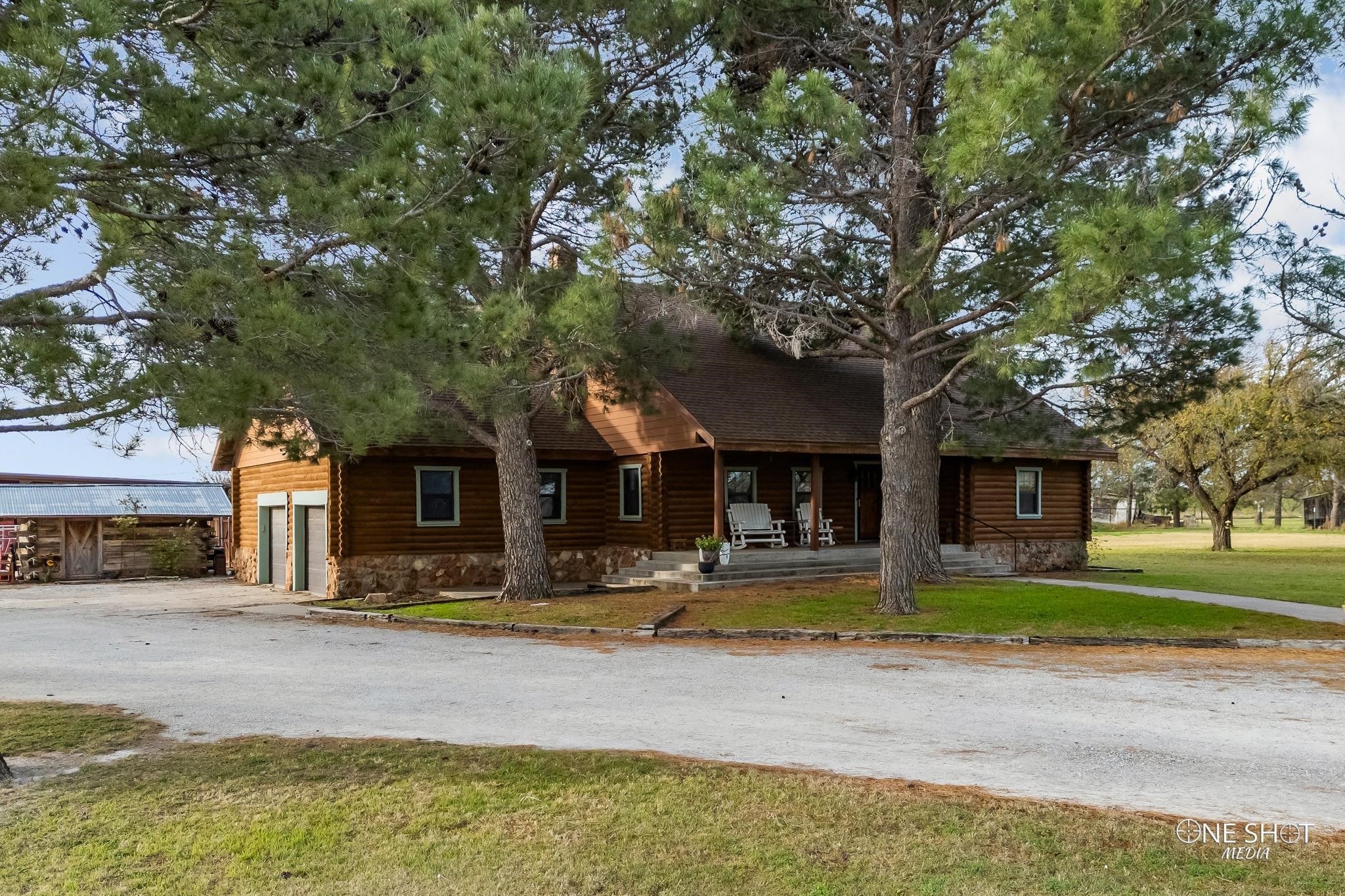 Log home with a stone foundation and covered porch, partly shaded by large pine trees.