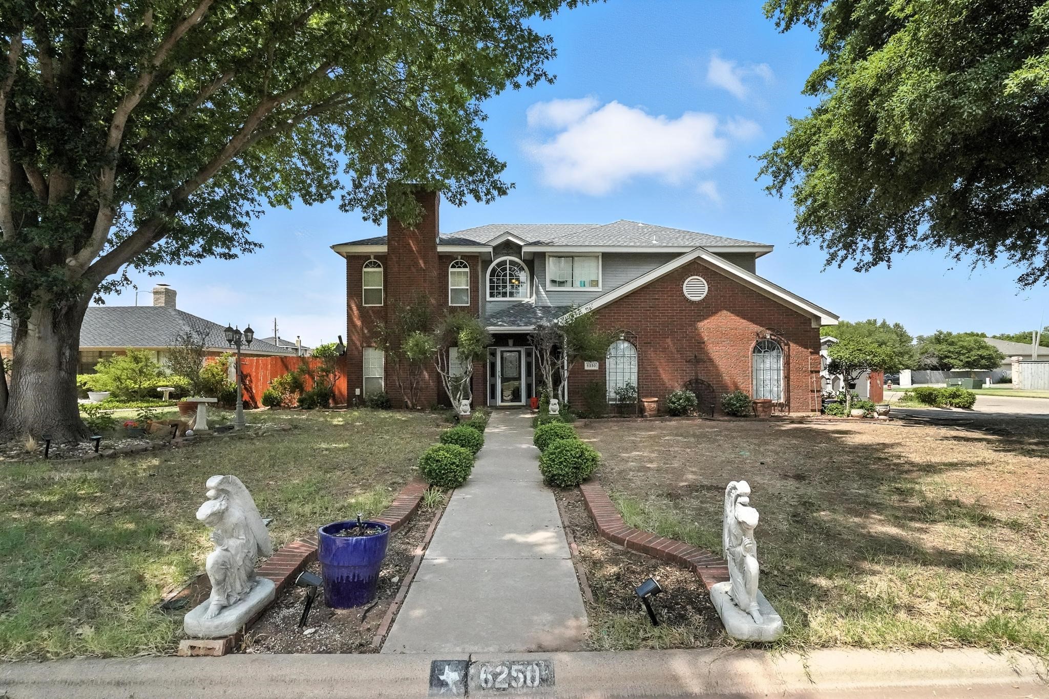 Two-story brick home with gray siding, mature trees, and a statue-lined front walkway.