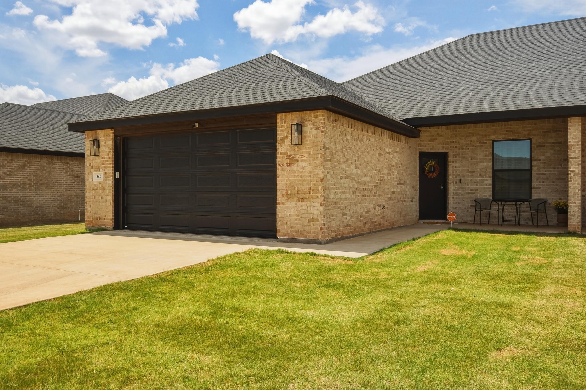Tan brick home with a dark double garage door, covered patio, and green front lawn.