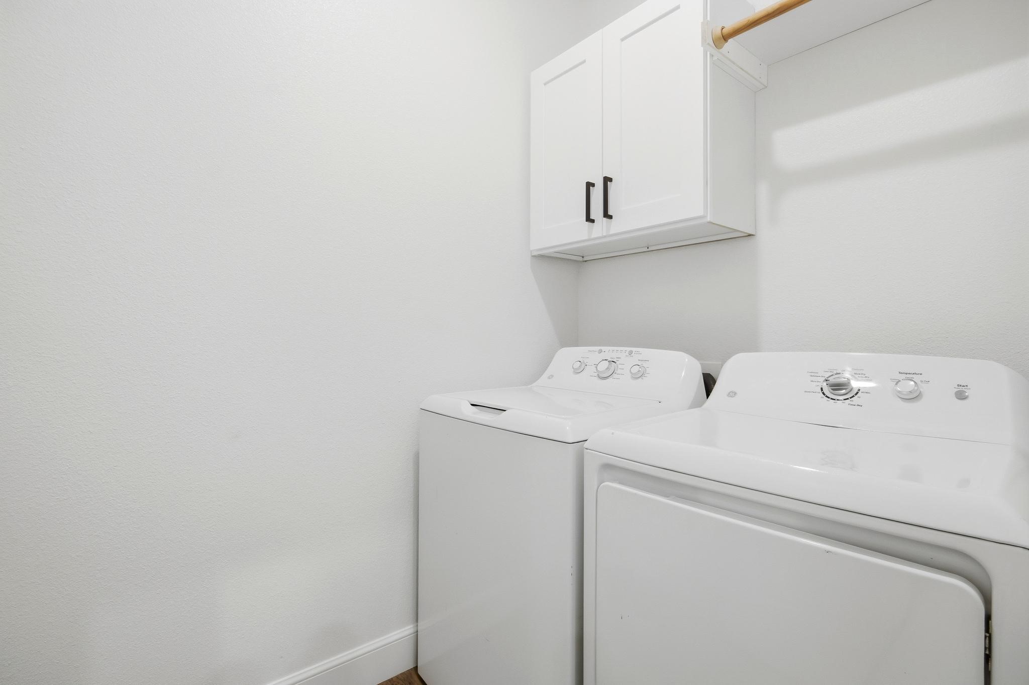 Laundry room with white washer and dryer beneath cabinets and a wooden hanging rod.