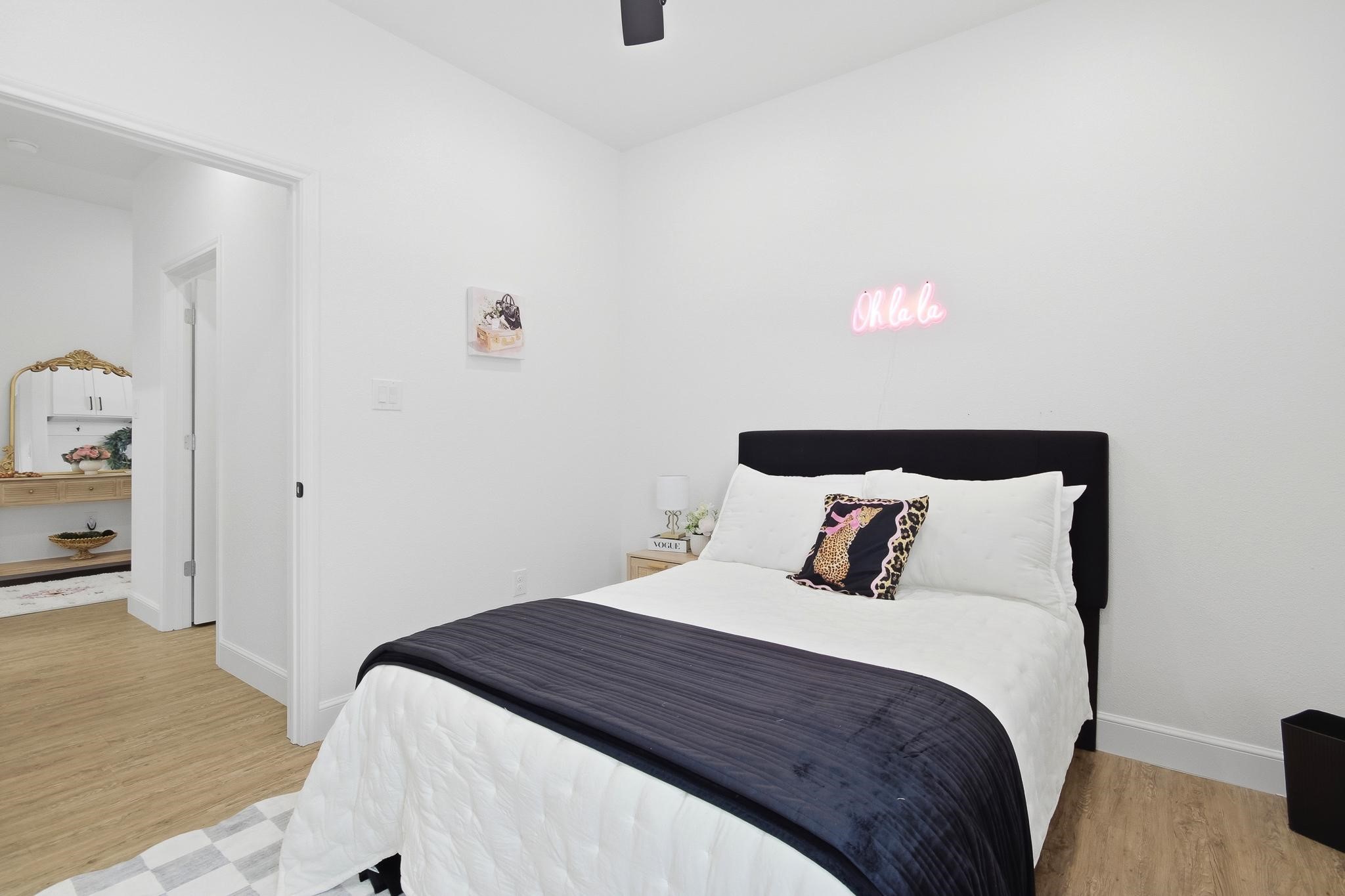 Staged bedroom with white bedding, a black headboard, and a pink “Oh la la” neon sign.