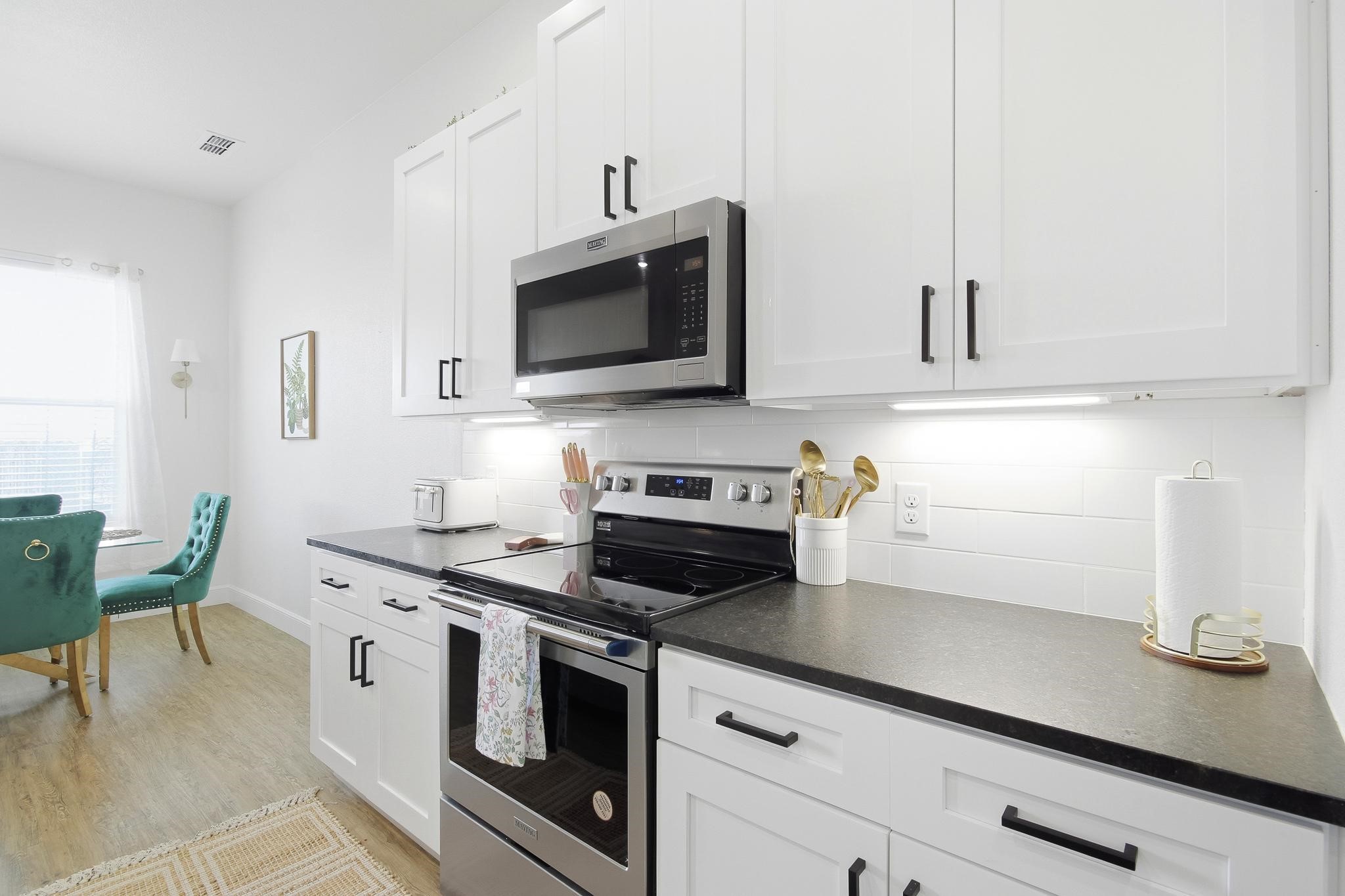 Bright kitchen with white cabinets, dark counters, stainless steel appliances, and teal chairs by a window.