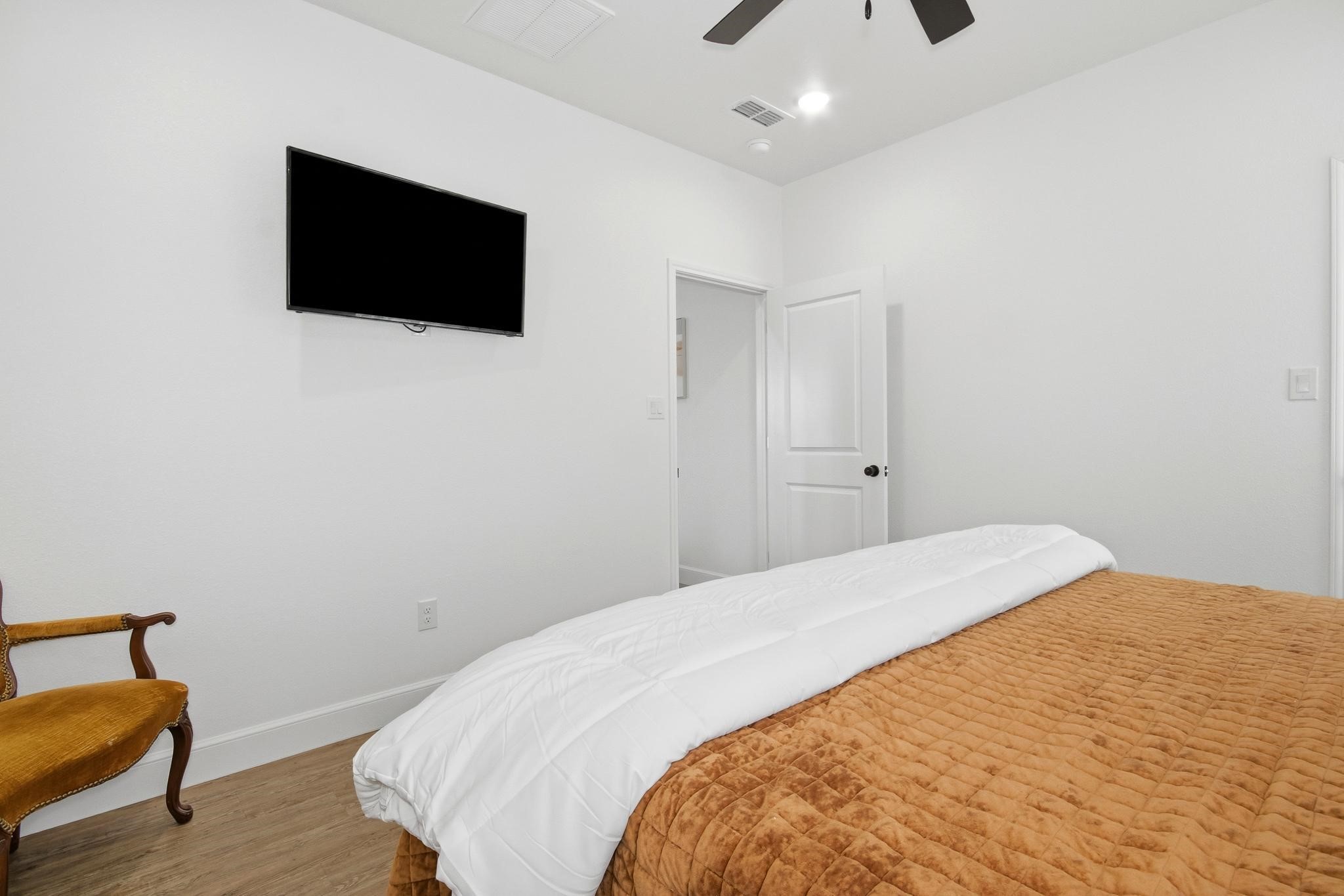 Bedroom with a rust-colored quilt, white bedding, wall-mounted TV, and wooden chair.