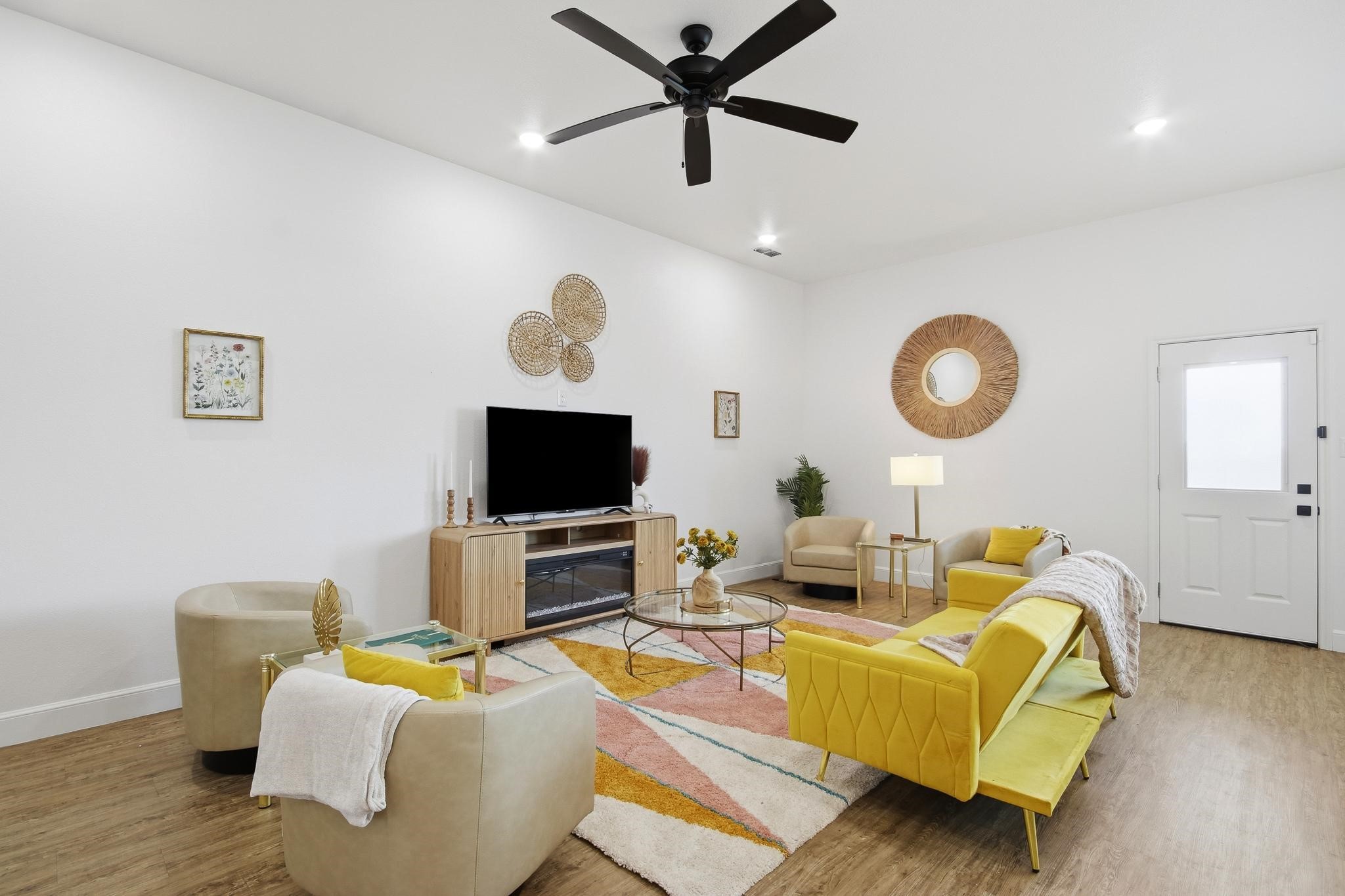 Staged living room with a yellow sofa, cream chairs, TV fireplace console, and colorful geometric rug.