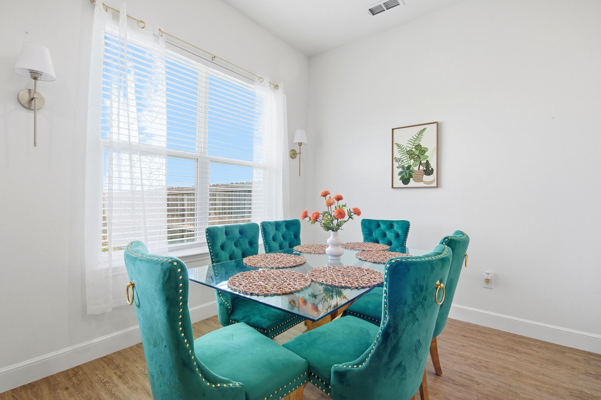 Staged dining room with a glass table, six teal velvet chairs, and a large window with white blinds.