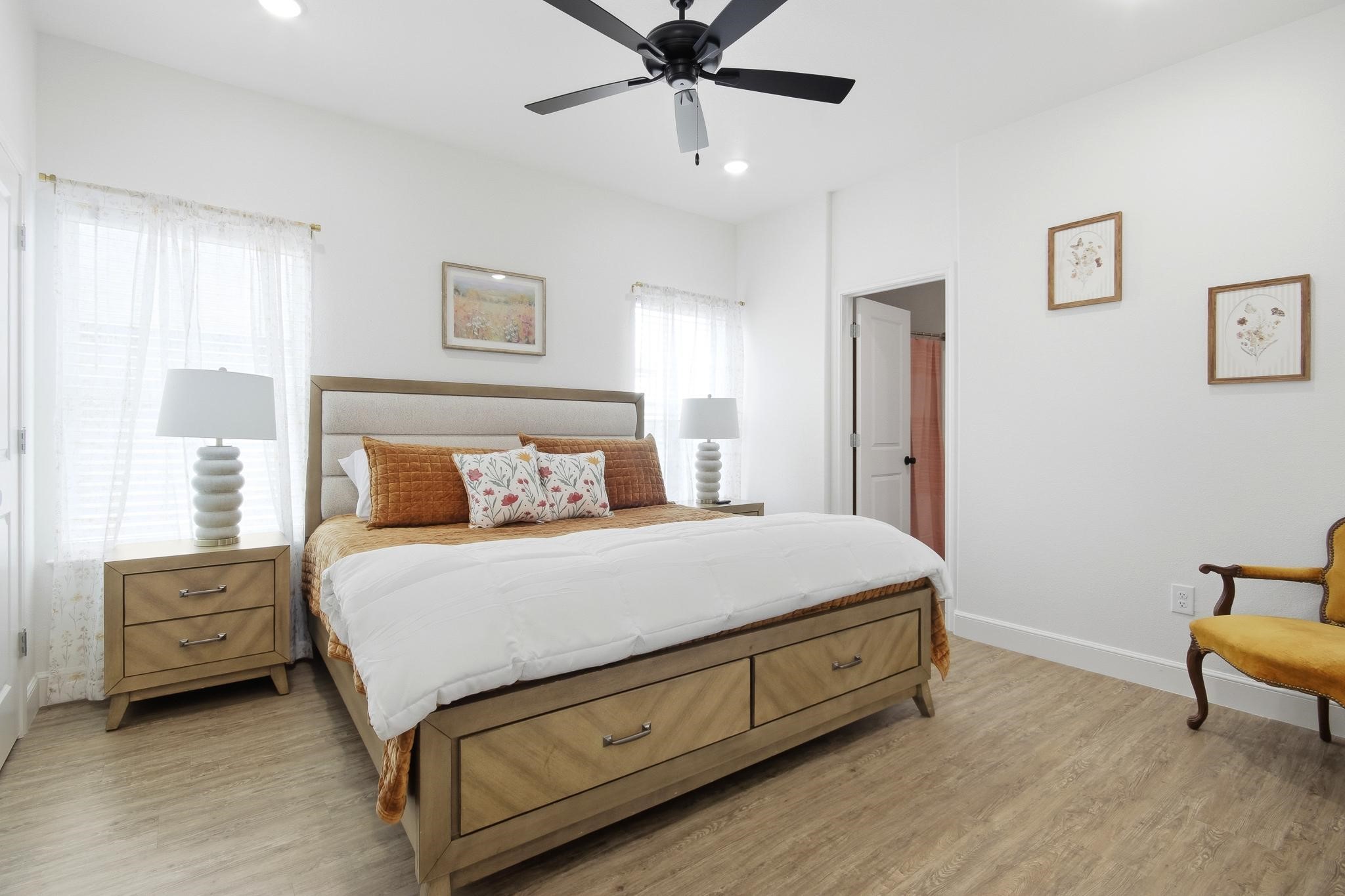 Staged bedroom with a wood storage bed, white bedding, rust pillows, and a black ceiling fan.