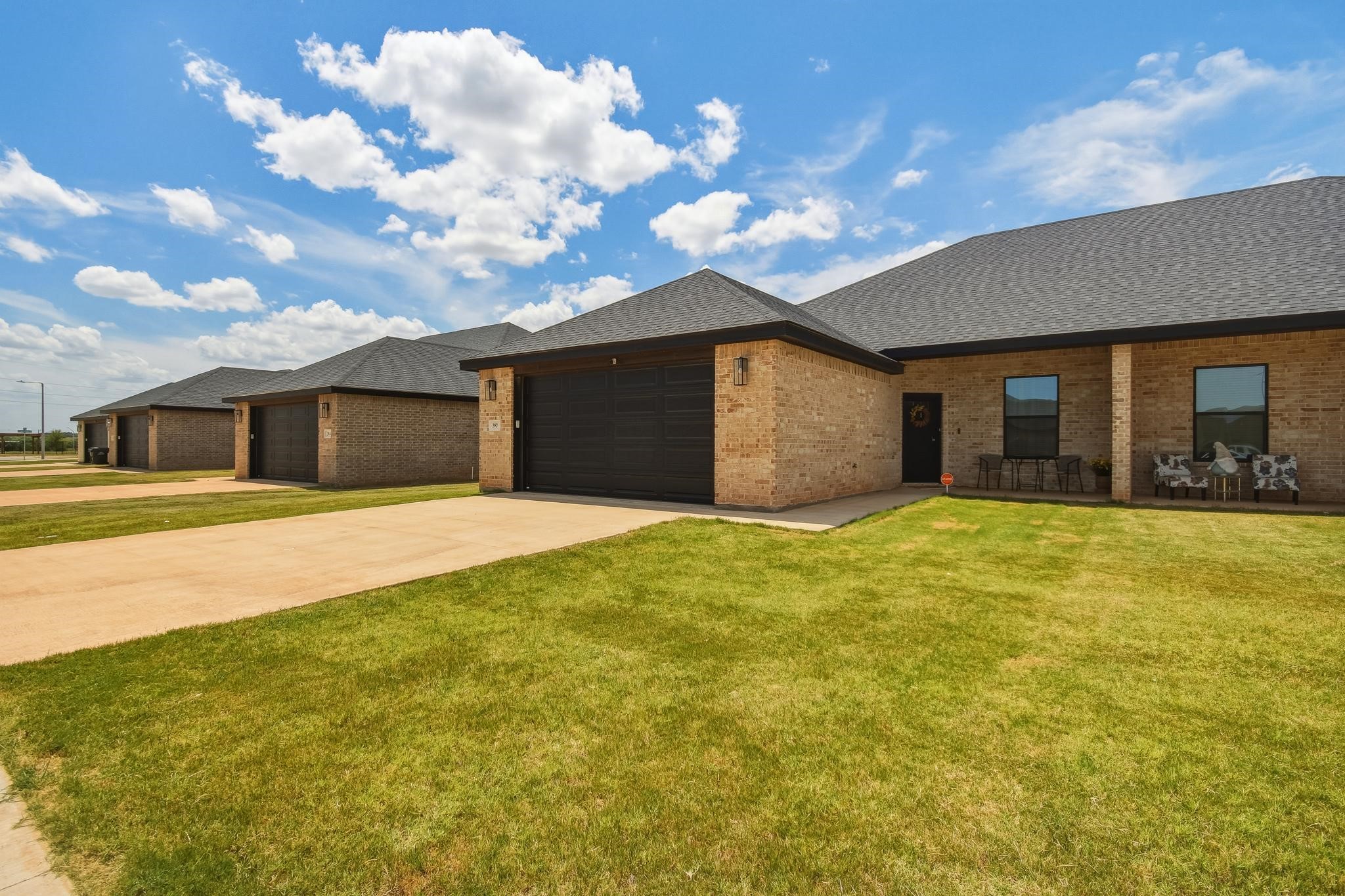 Brick homes with dark garage doors, broad driveways, green lawns, and patio seating.