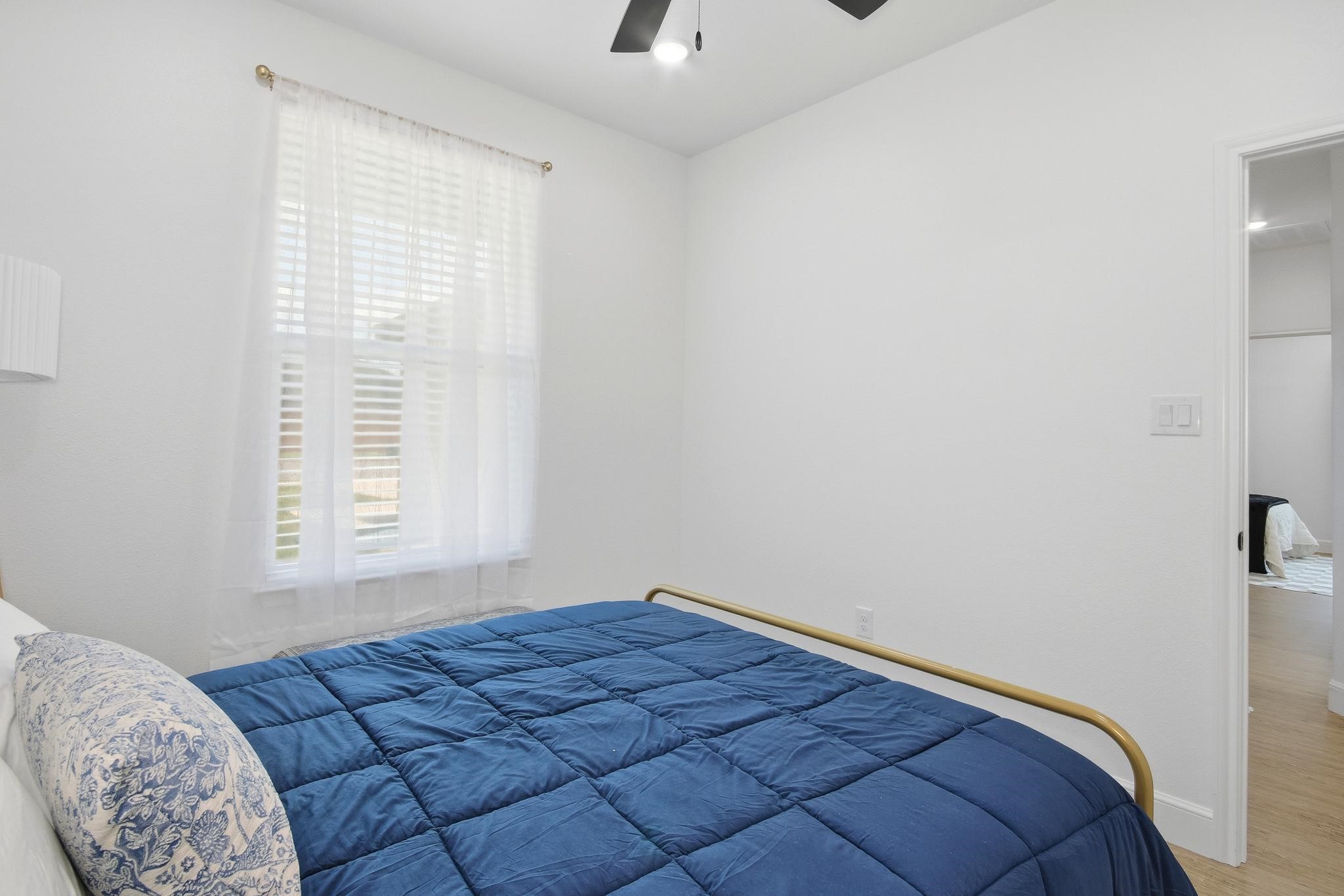 Bedroom with a deep blue quilted bed, sheer-curtained window, and doorway to another room.