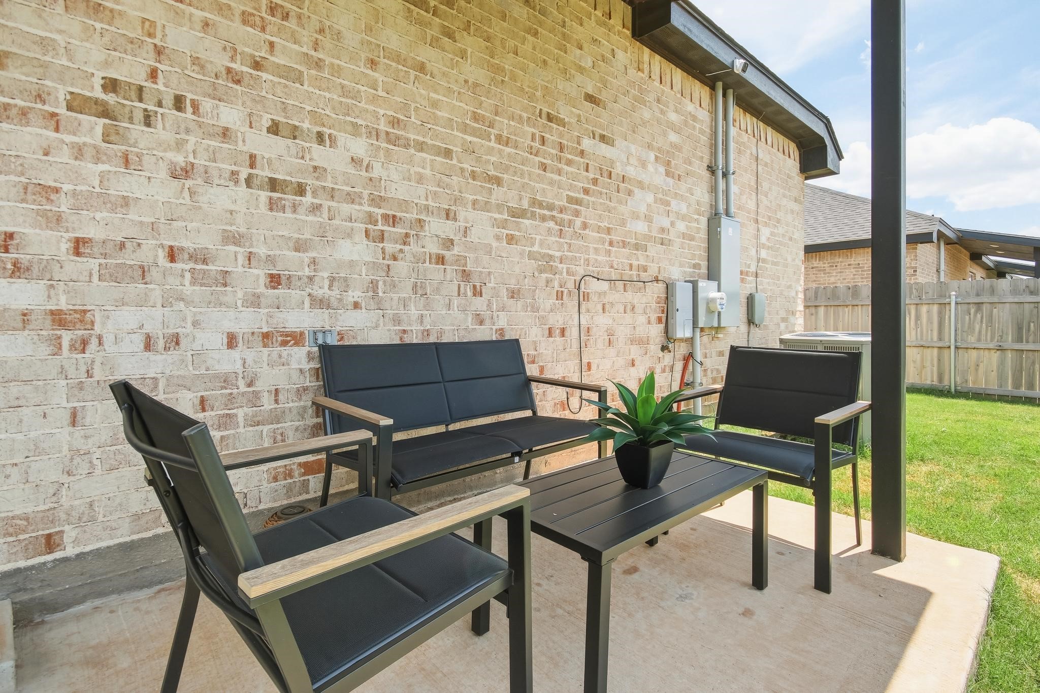 Covered brick patio furnished with black seating and a coffee table beside a fenced lawn.