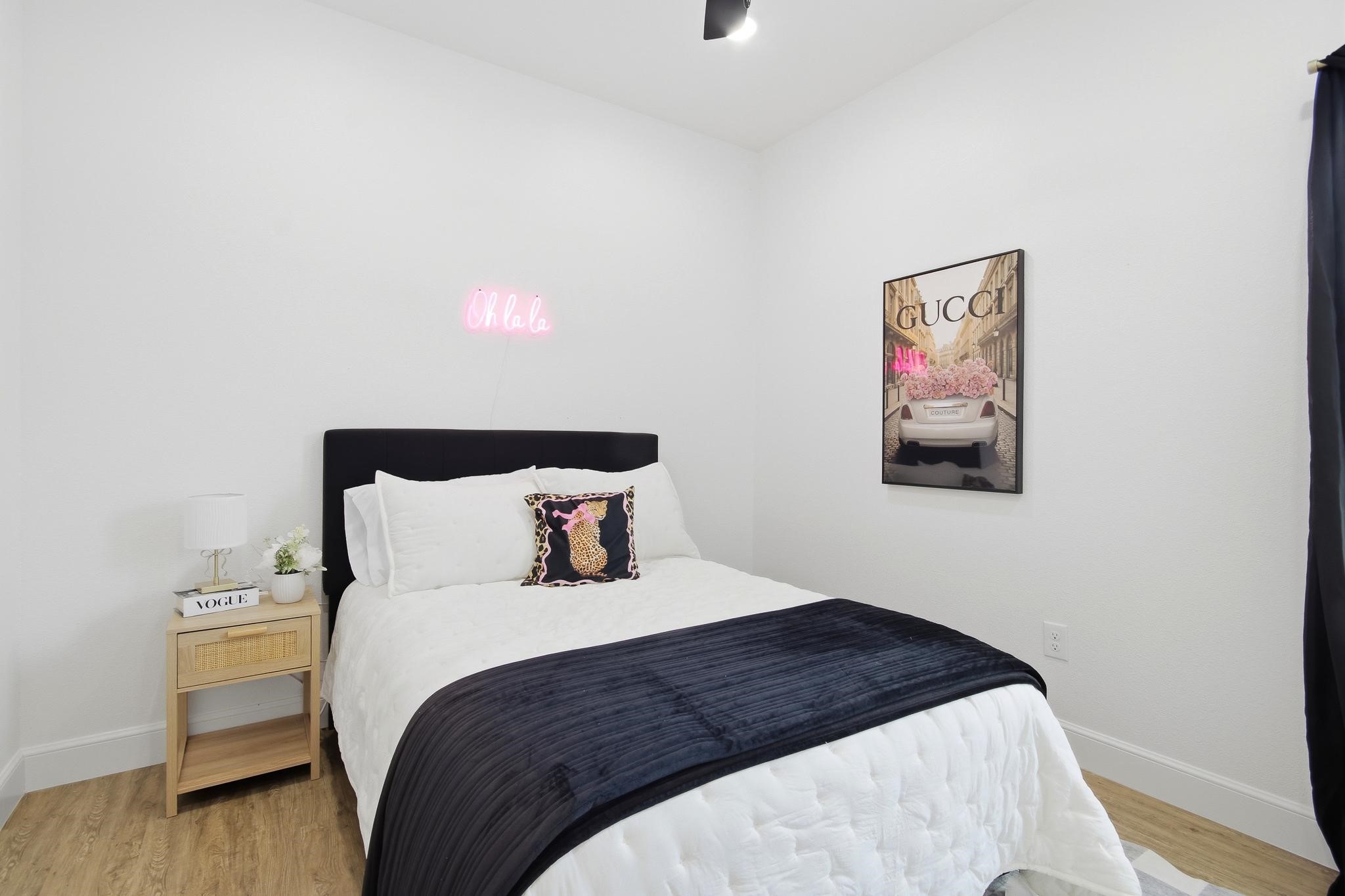 Bedroom with a white bed, navy throw, pink neon sign, and framed fashion artwork.