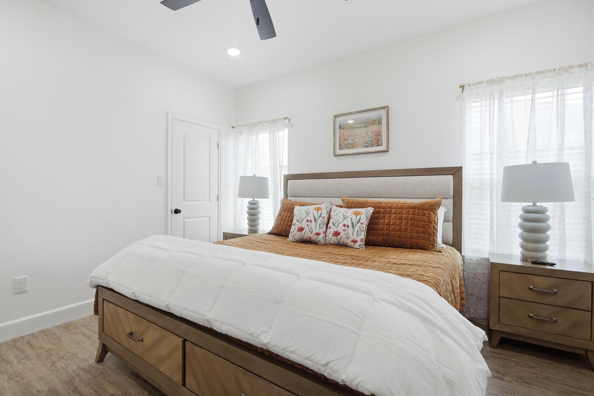 Bedroom with a wood-framed bed, rust-colored pillows, matching nightstands, and sheer-curtained windows.