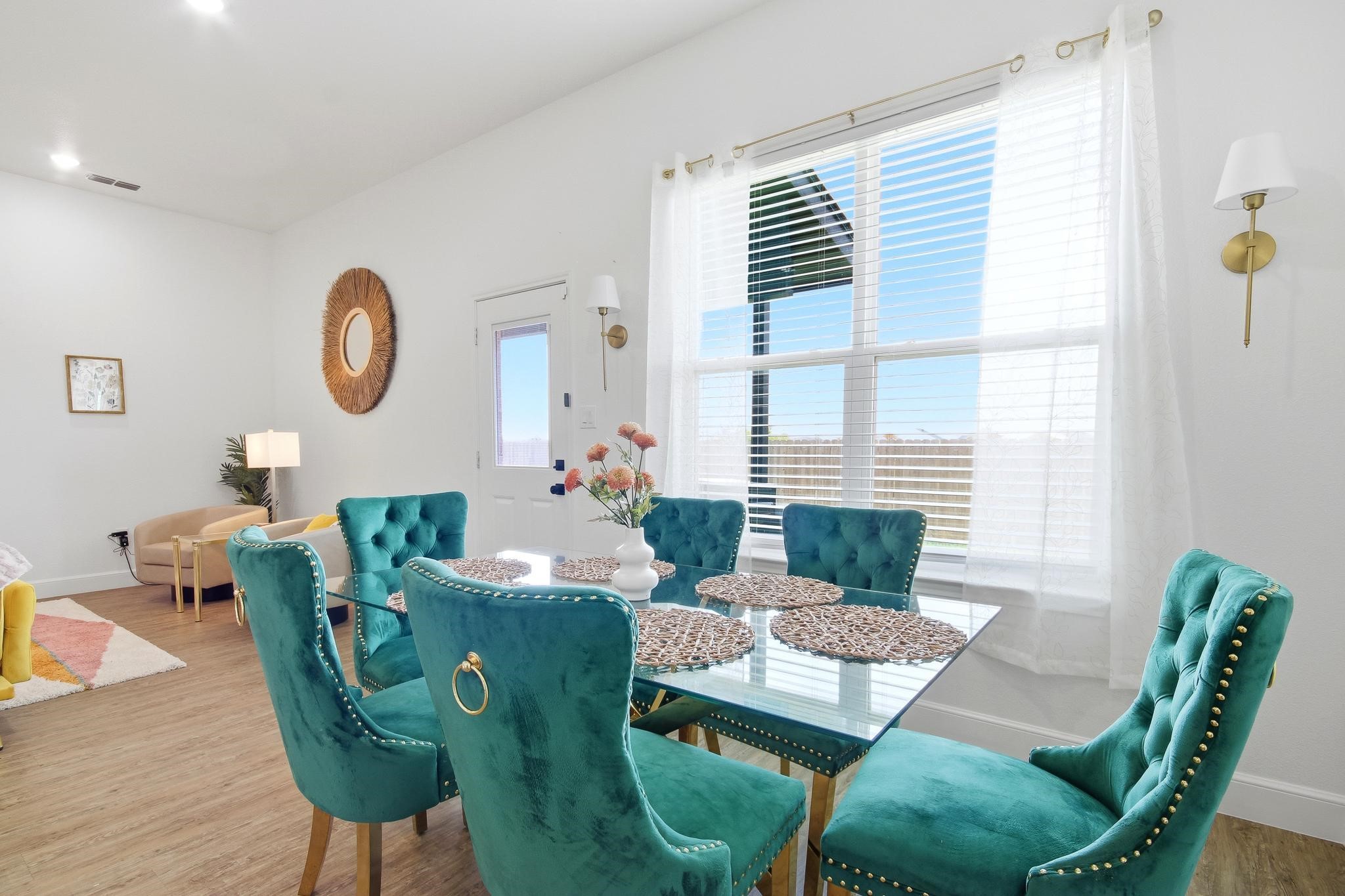 Dining room with a glass table, teal tufted chairs, and a large bright window.