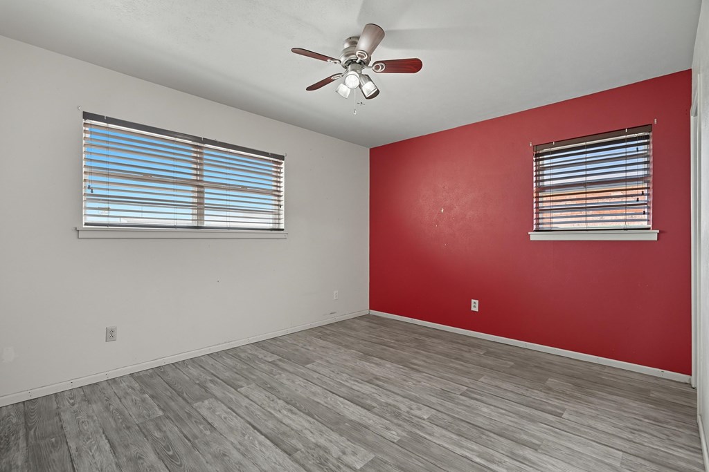 Empty room with gray plank flooring, a red accent wall, two windows, and a ceiling fan.