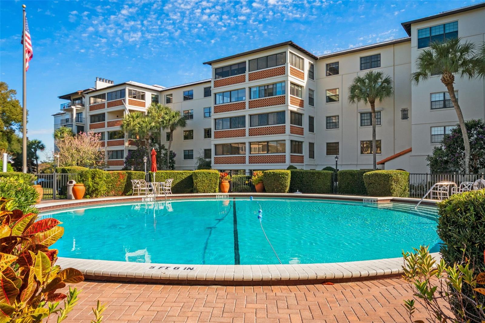 Outdoor pool in front of a condominium building, with tropical landscaping and patio seating.