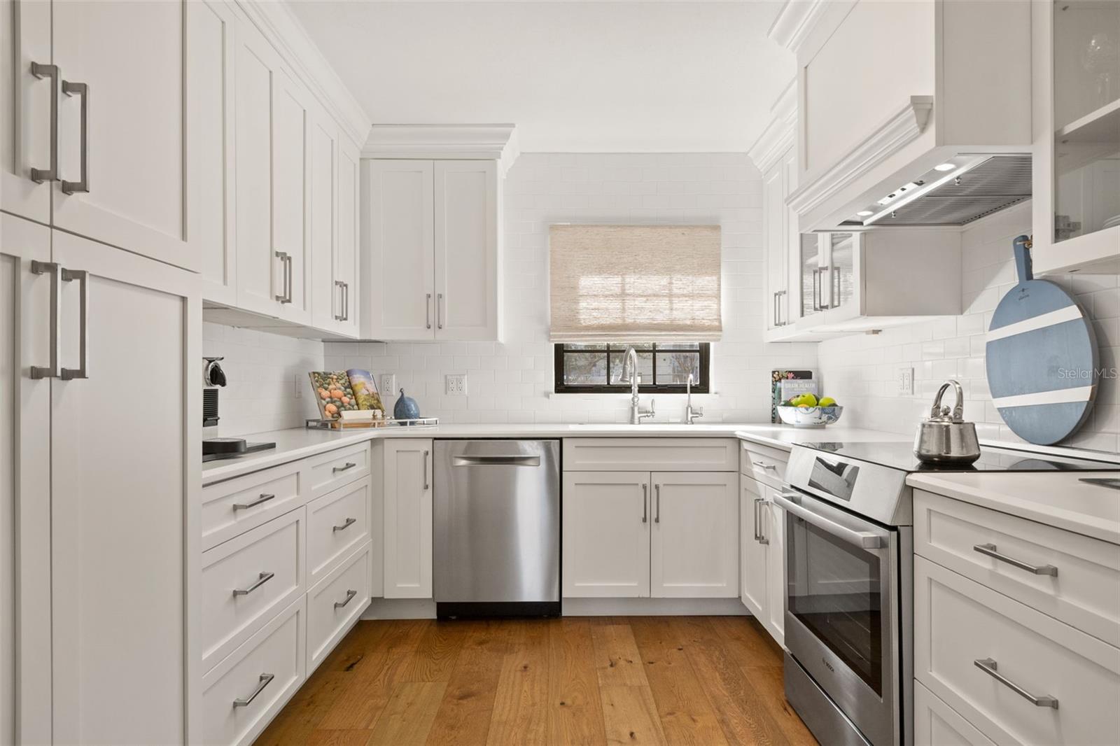 Bright white kitchen with stainless steel appliances, wood floors, and a window above the sink.