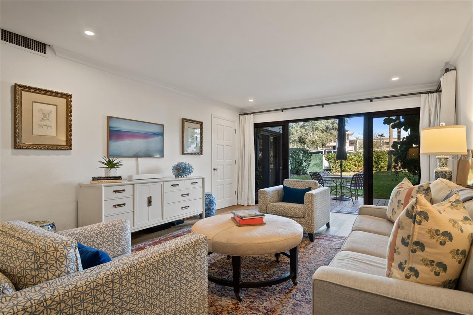Furnished living room with patterned chairs, a round ottoman, and sliding doors opening to a garden.
