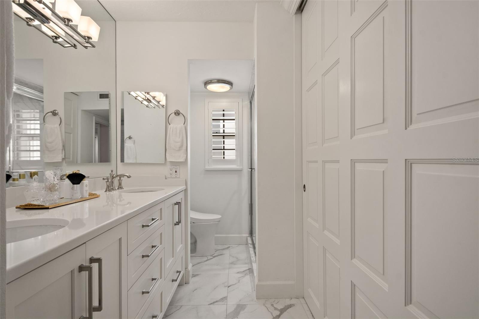 Bathroom with a white double-sink vanity, marble-look floors, shuttered window, and glass shower.