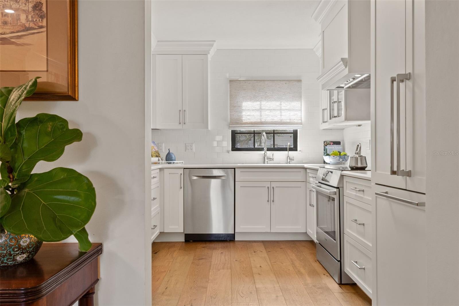 White kitchen with stainless steel appliances, a window above the sink, and light wood floors.