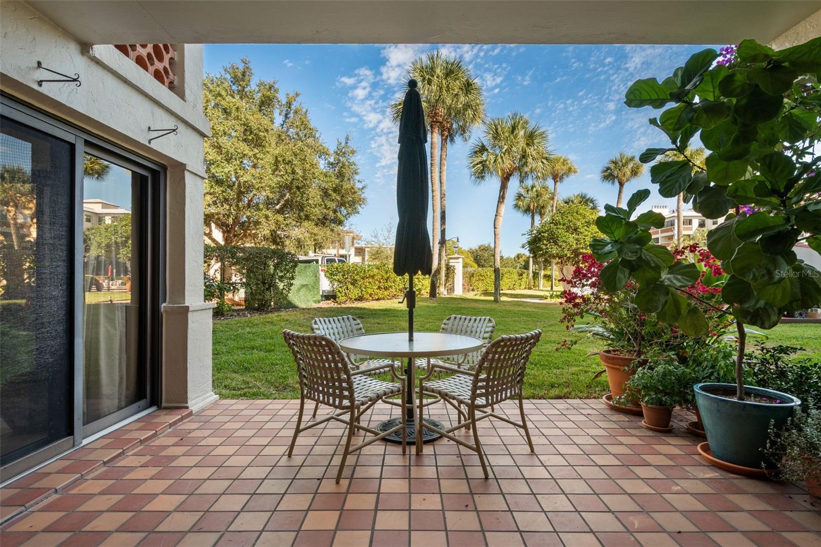 Tiled patio with an outdoor dining set, closed umbrella, and potted plants overlooking a palm-lined lawn.
