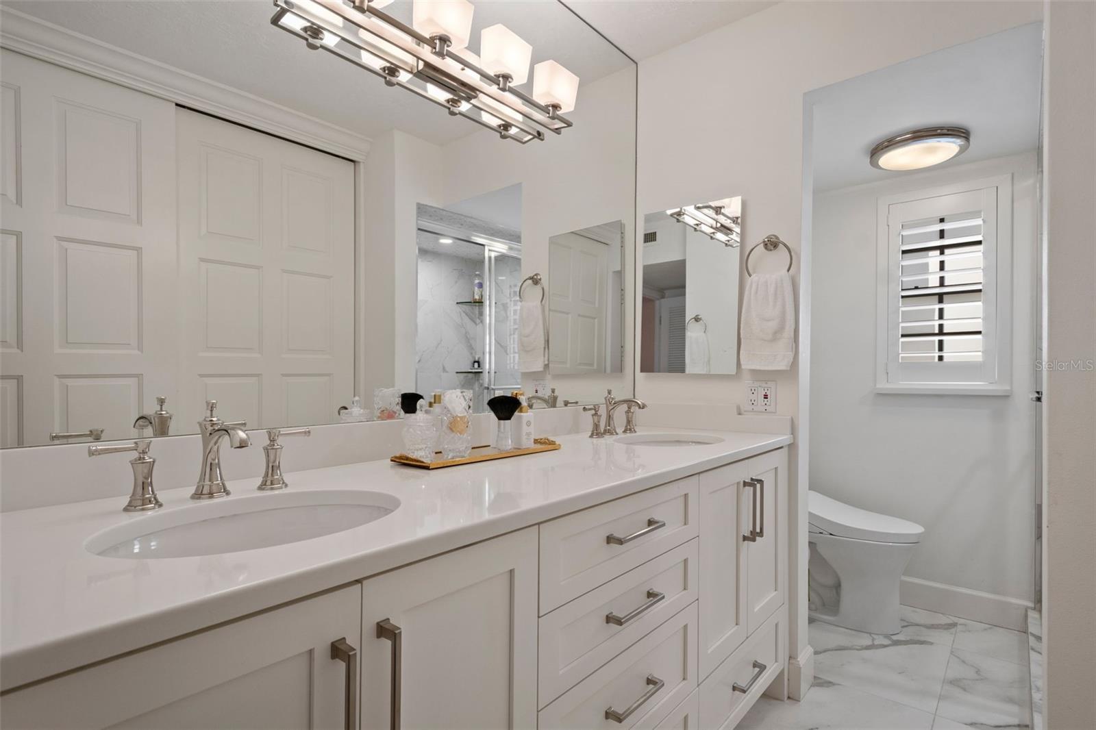 Bright bathroom with a long double vanity, paneled white cabinets, marble-look flooring, and plantation shutters.