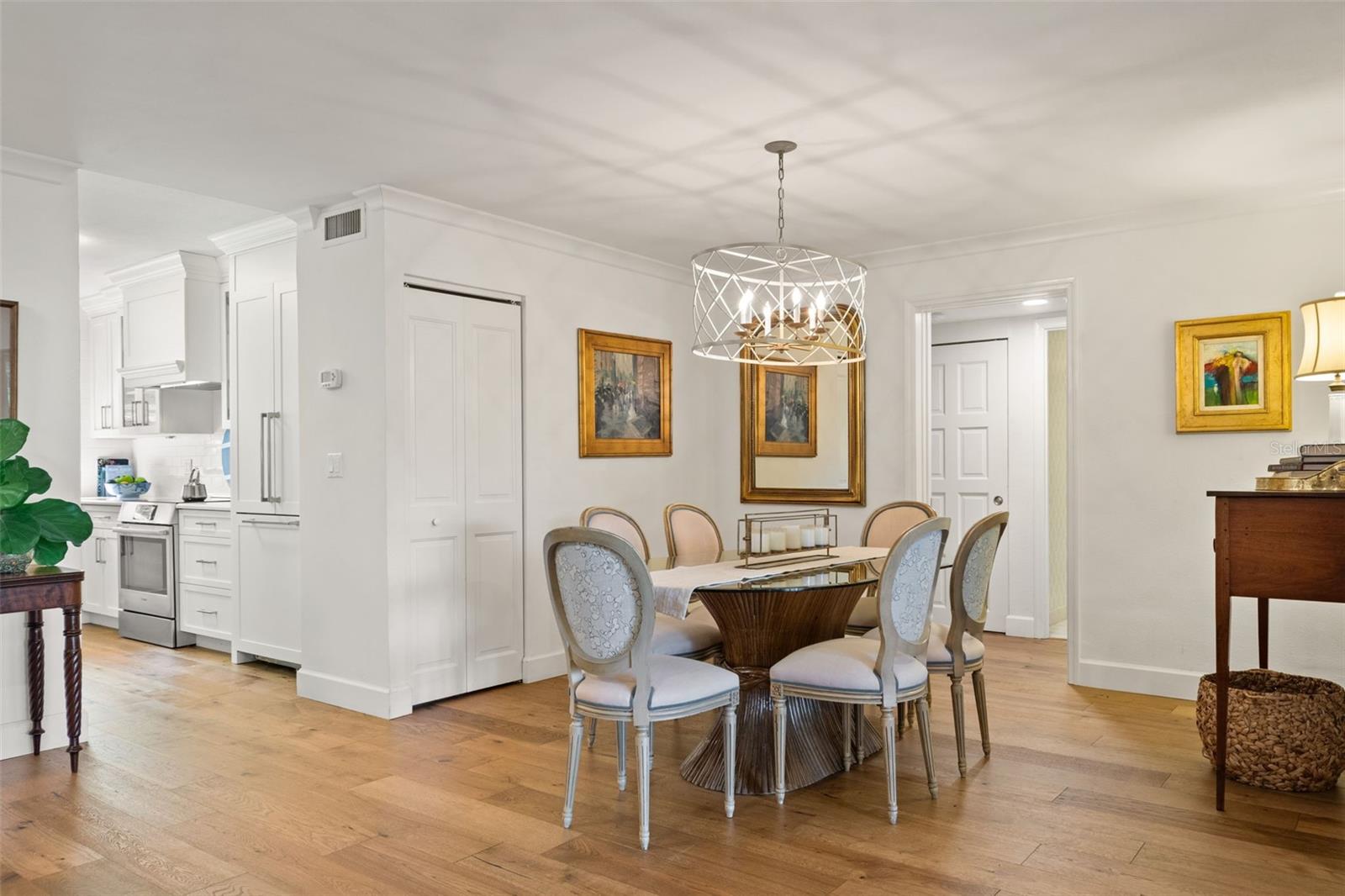 Furnished dining area with a round wooden table, geometric pendant light, and adjoining white kitchen.