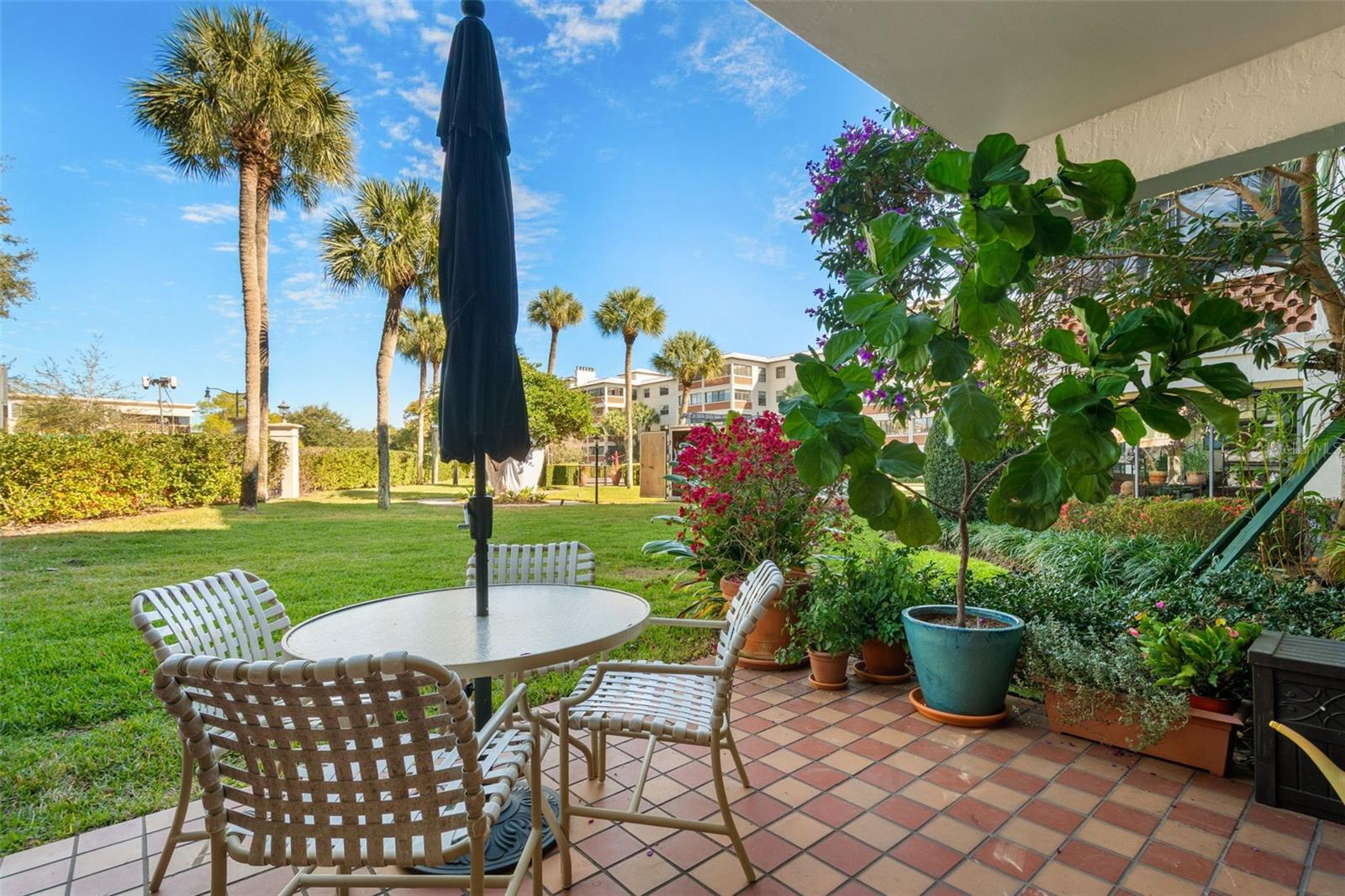 Condominium patio with tiled floor, white seating, closed umbrella, and landscaped lawn with palm trees.