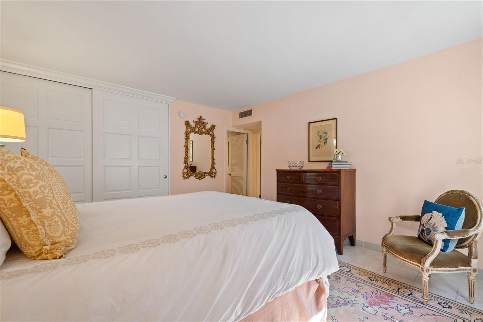Bedroom with a white bed, paneled closet doors, dark dresser, ornate mirror, and patterned rug.