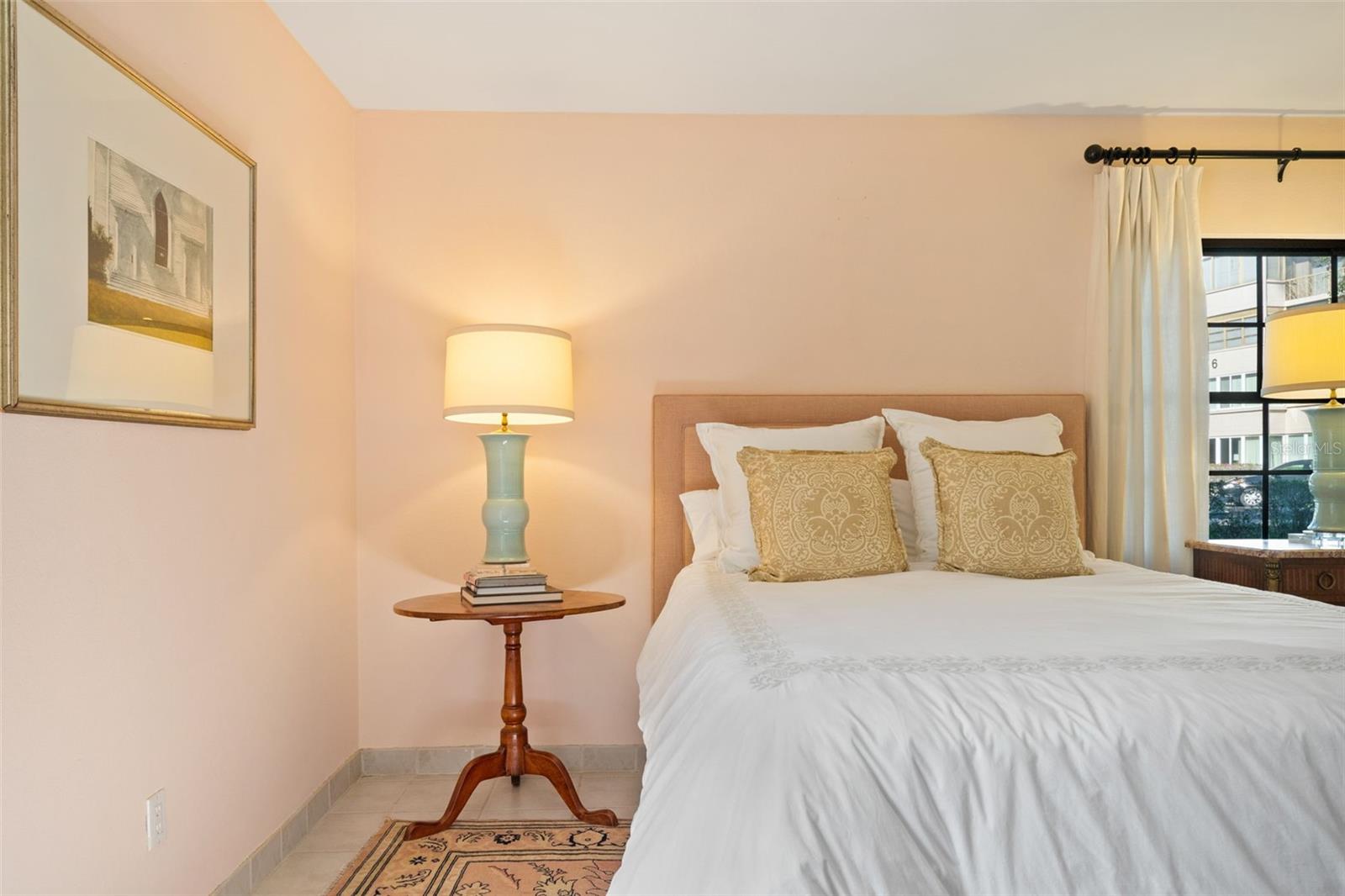 Bedroom with a white bed, gold patterned pillows, and a turquoise lamp beside the window.