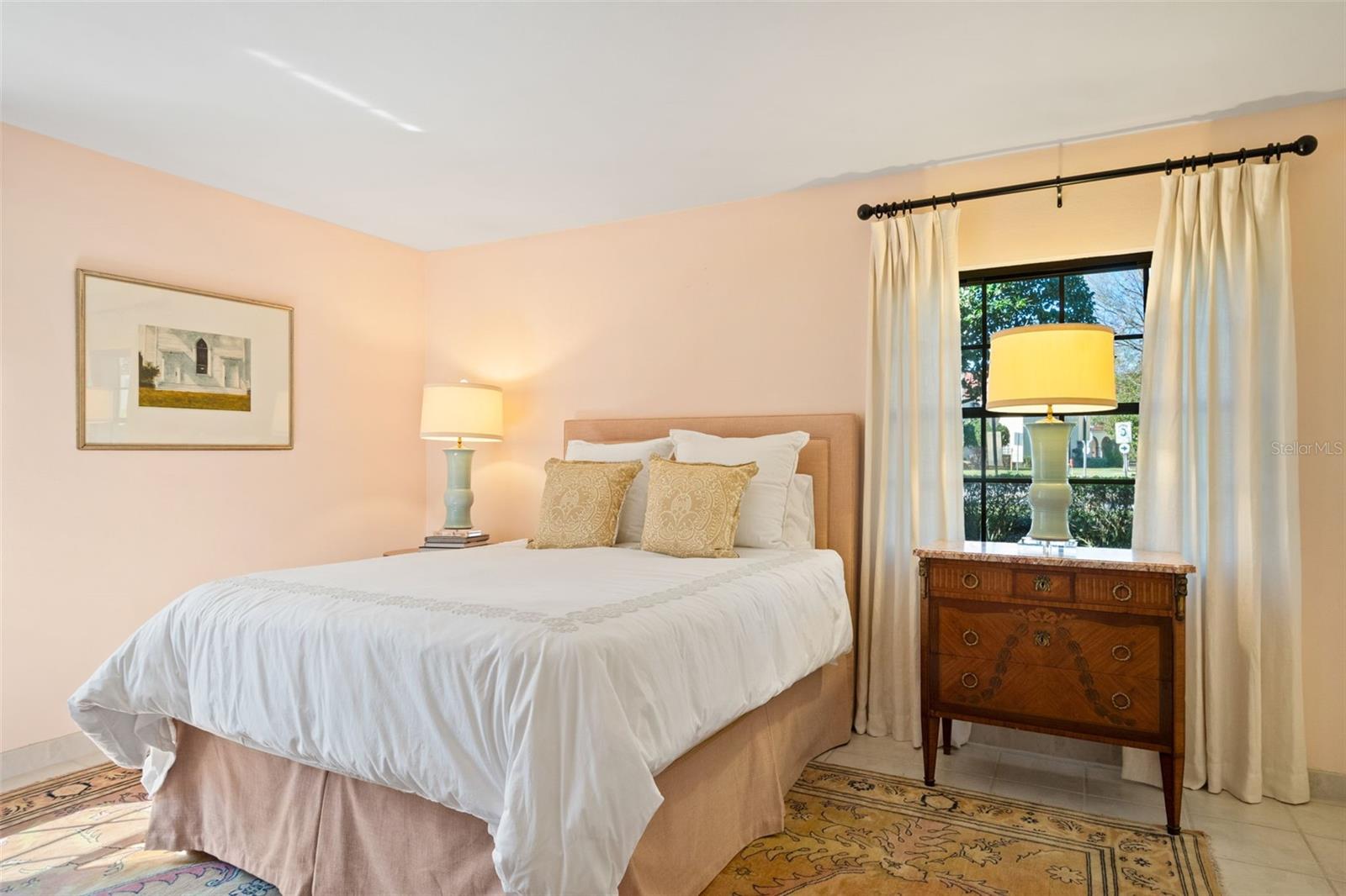 Furnished bedroom with a white bed, patterned rug, and wooden dresser by a curtained window.