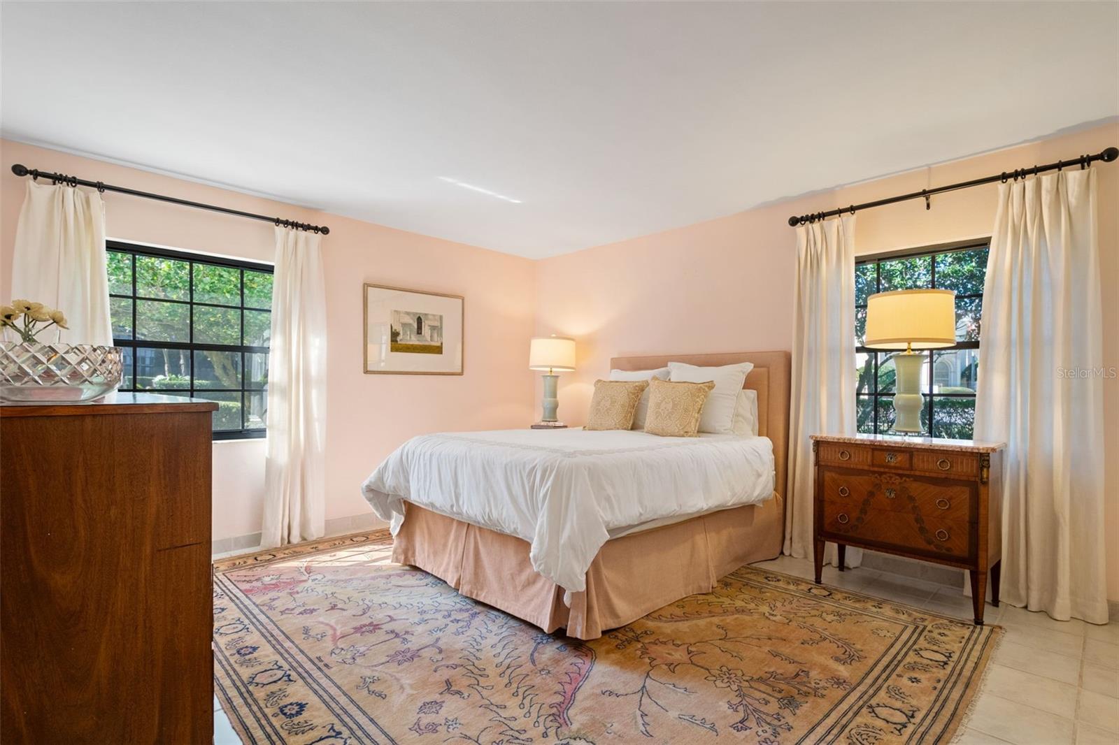 Bedroom with a king-sized bed, peach walls, two curtained windows, and a patterned rug.
